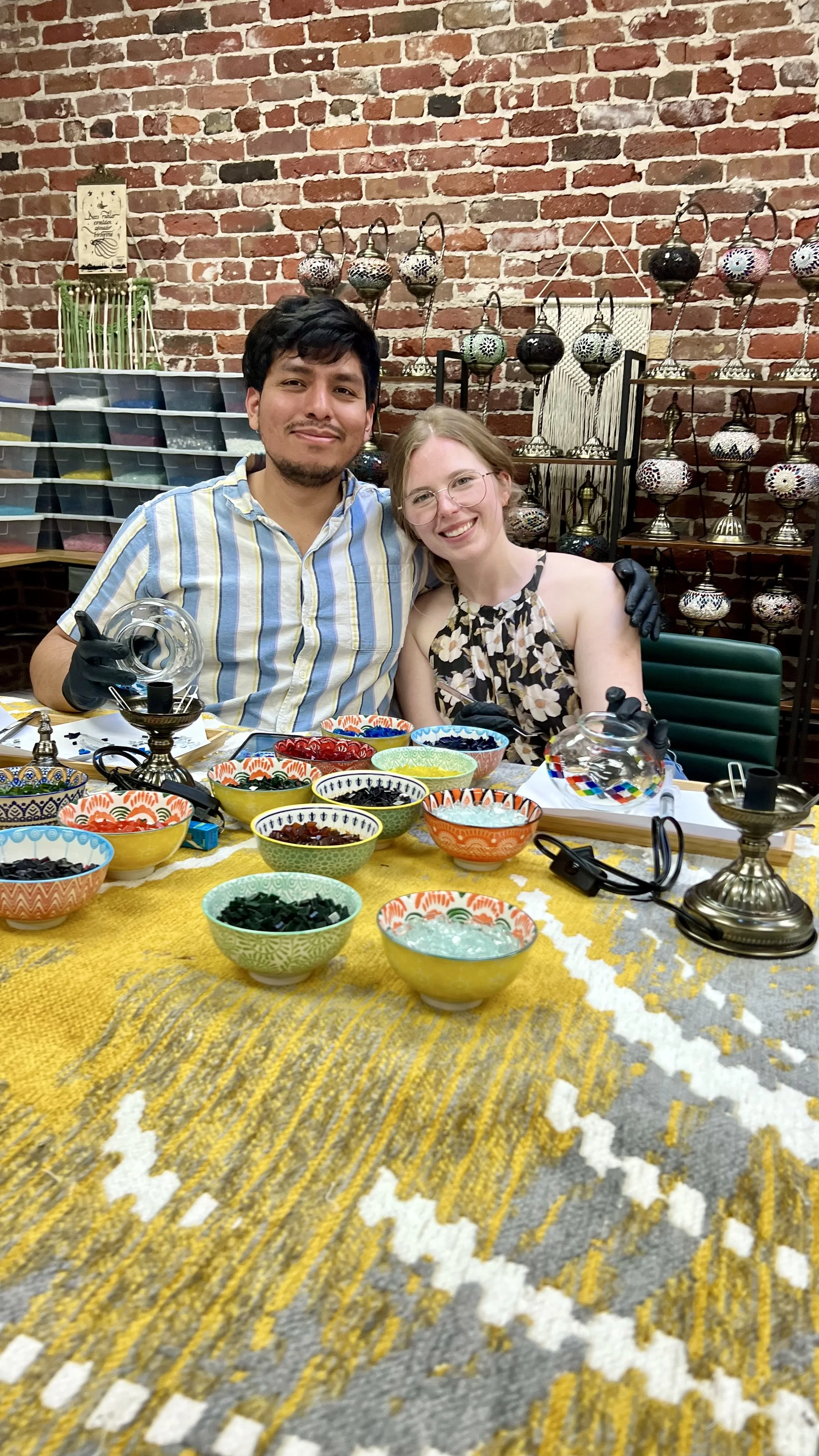 Two people sitting at a table with colorful bowls and a brick wall background, smiling for a photo.