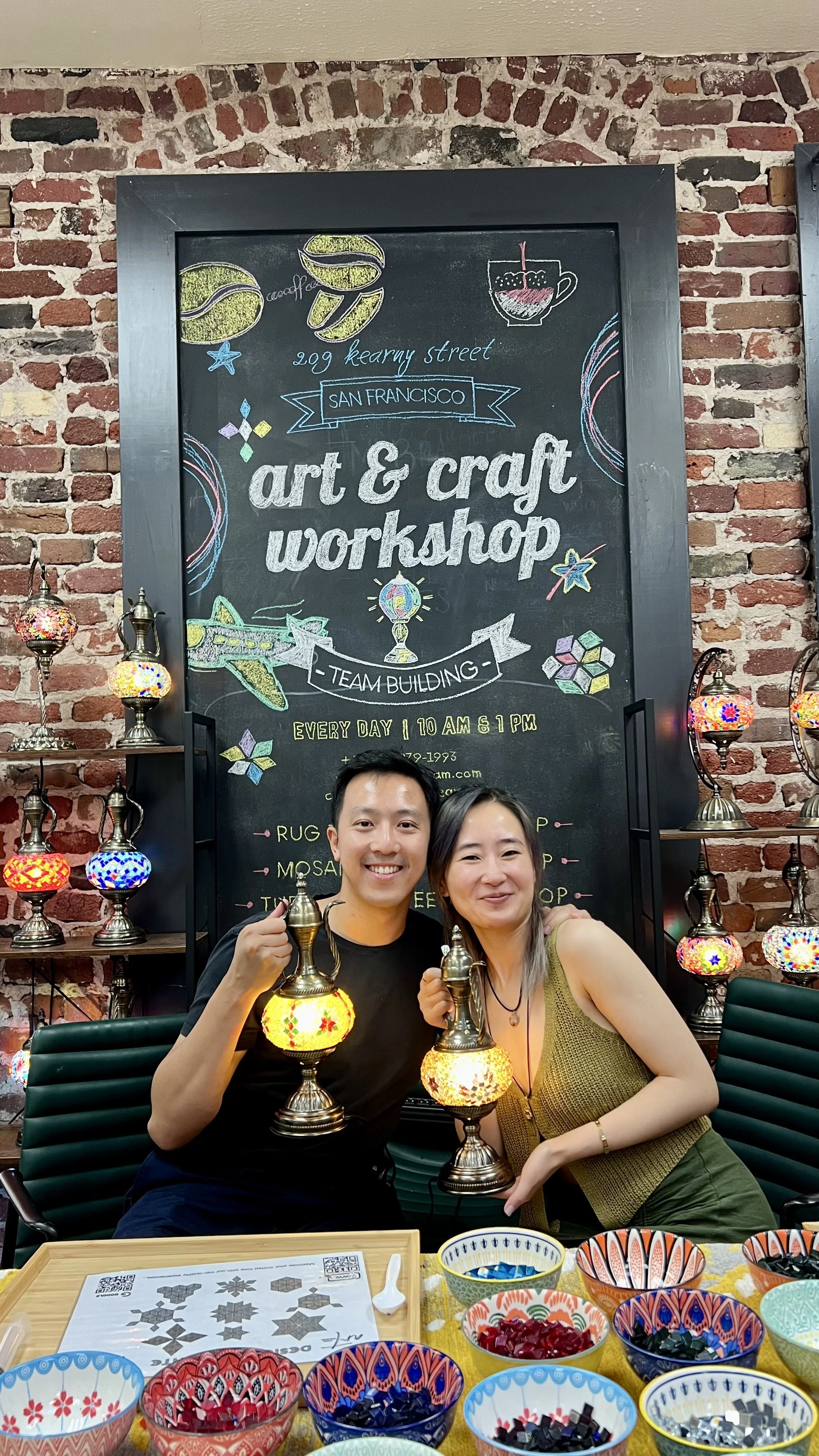 Two smiling people holding lamps at an art and craft workshop in front of a blackboard, with colorful bowls of craft supplies on the table and decorative lamps on shelves.