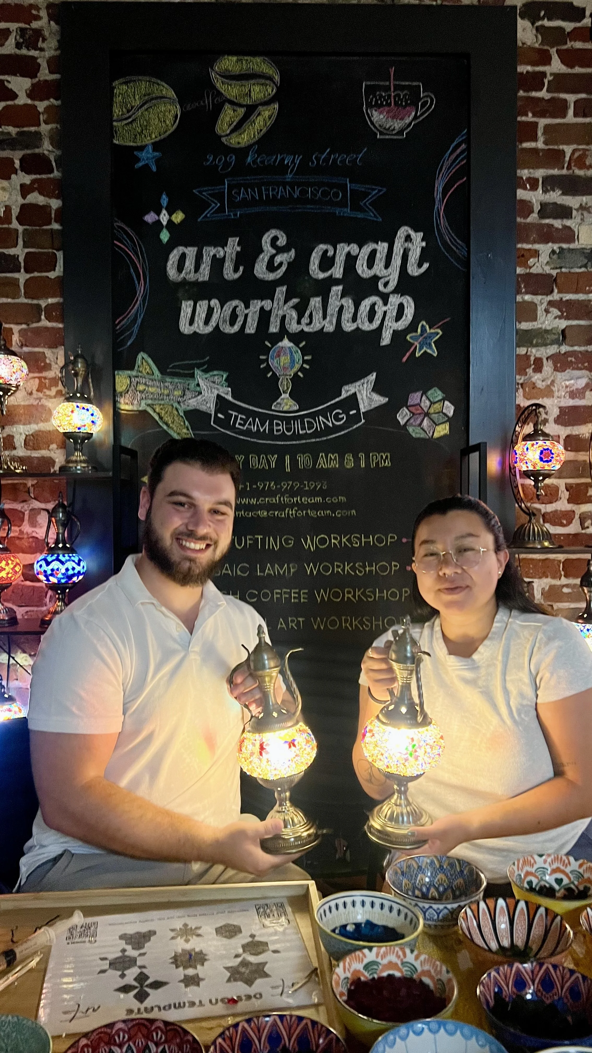 Two people holding decorative lamps in front of an art and craft workshop sign, with colorful bowls on the table.
