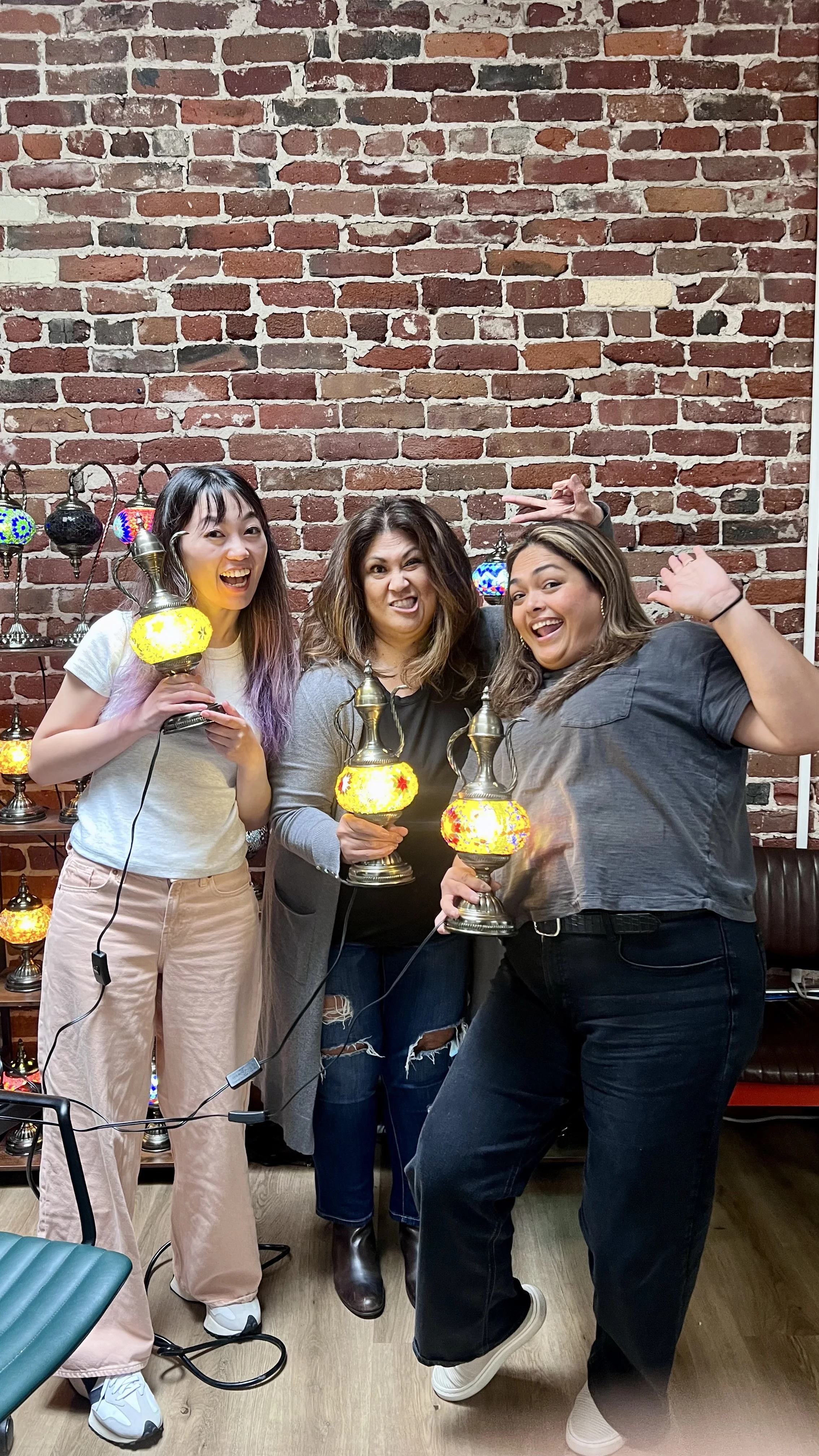 Three women standing in front of a brick wall, holding colorful Moroccan-style lamps, smiling and making playful faces.