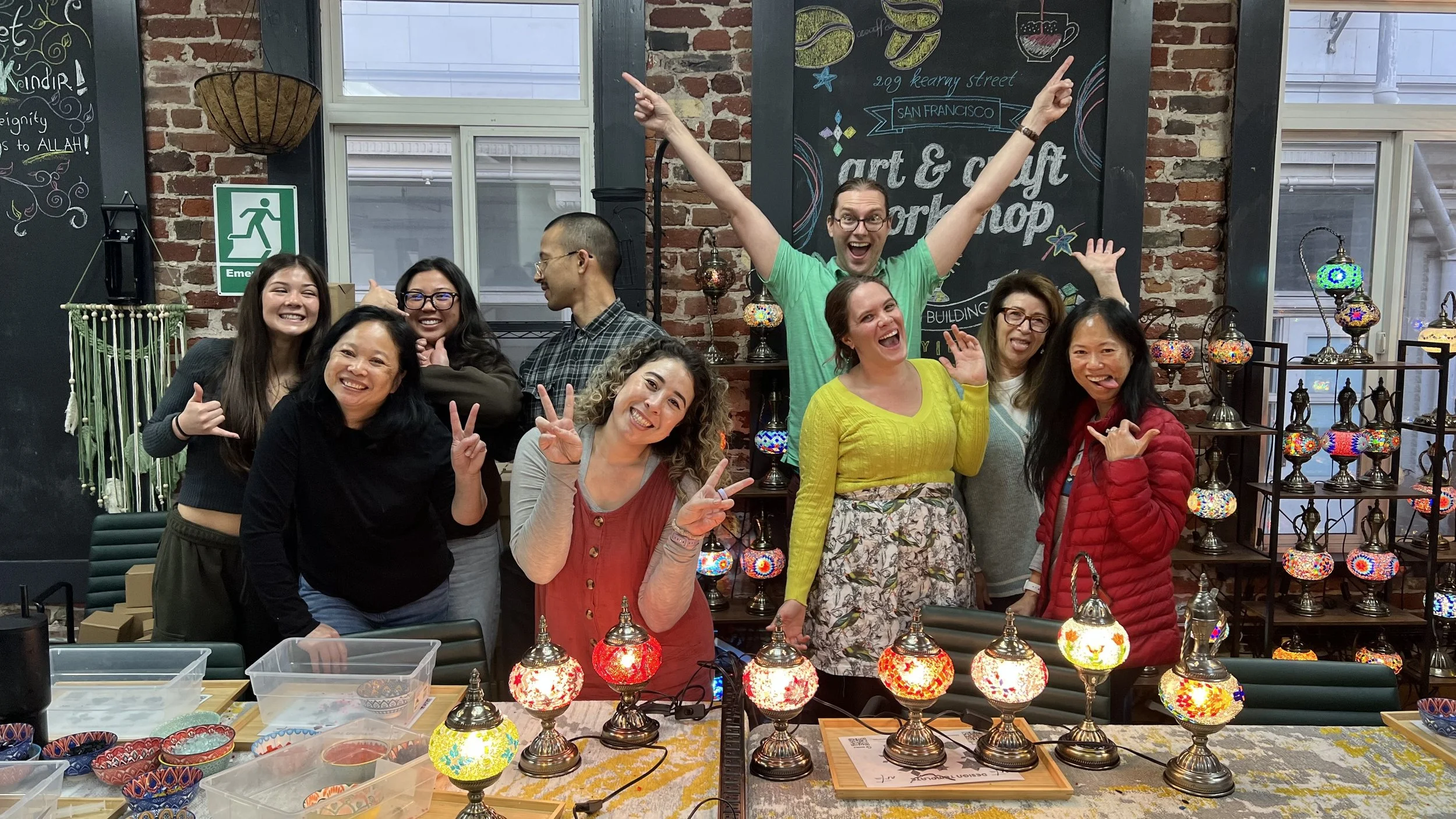 Group of nine people smiling and posing happily in a craft workshop, with colorful mosaic lamps on the table in front, brick wall background, and decorative items around.