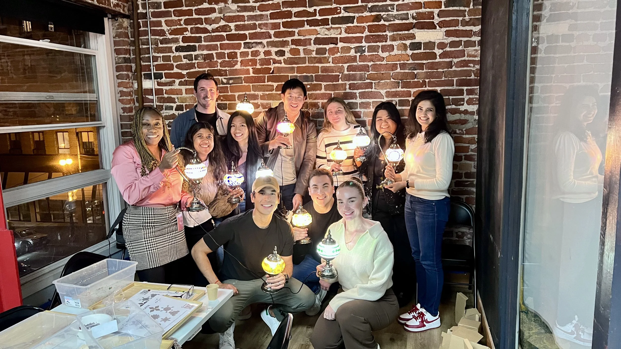 Group of people at a celebration holding decorative lamps, standing in a room with brick walls and large windows
