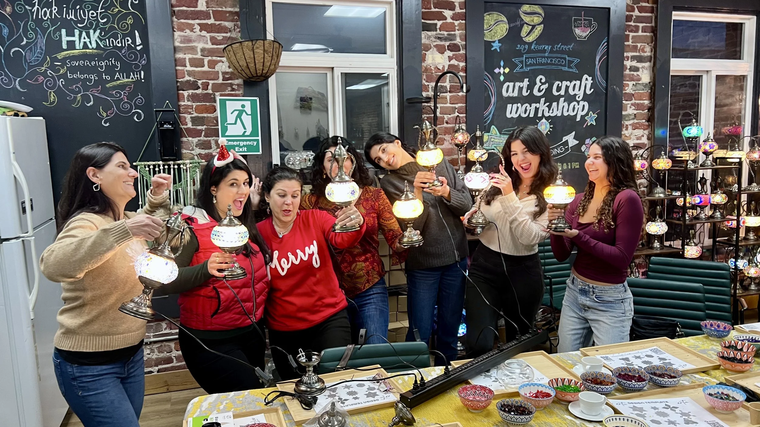 Seven women celebrating at a craft workshop, holding illuminated decorative lamps, with colorful bowls on the table and a blackboard with art and craft workshop signs in the background.