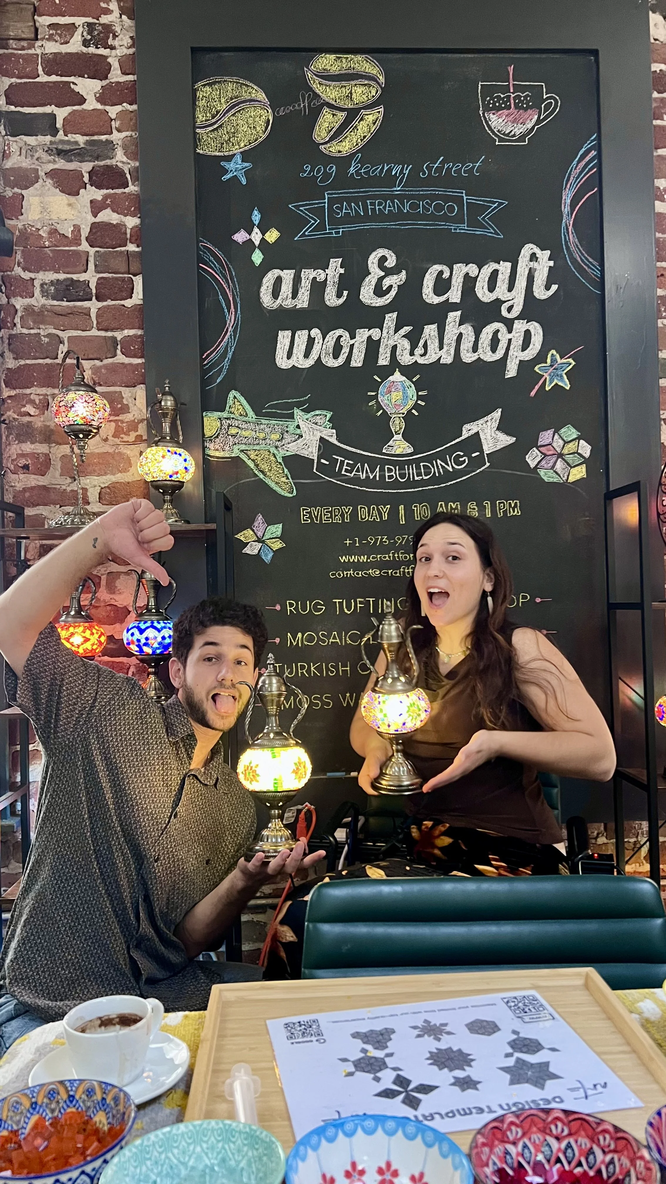 Two people celebrating at an art and craft workshop, holding colorful lanterns, sitting at a table with craft supplies, in front of a blackboard with workshop details.