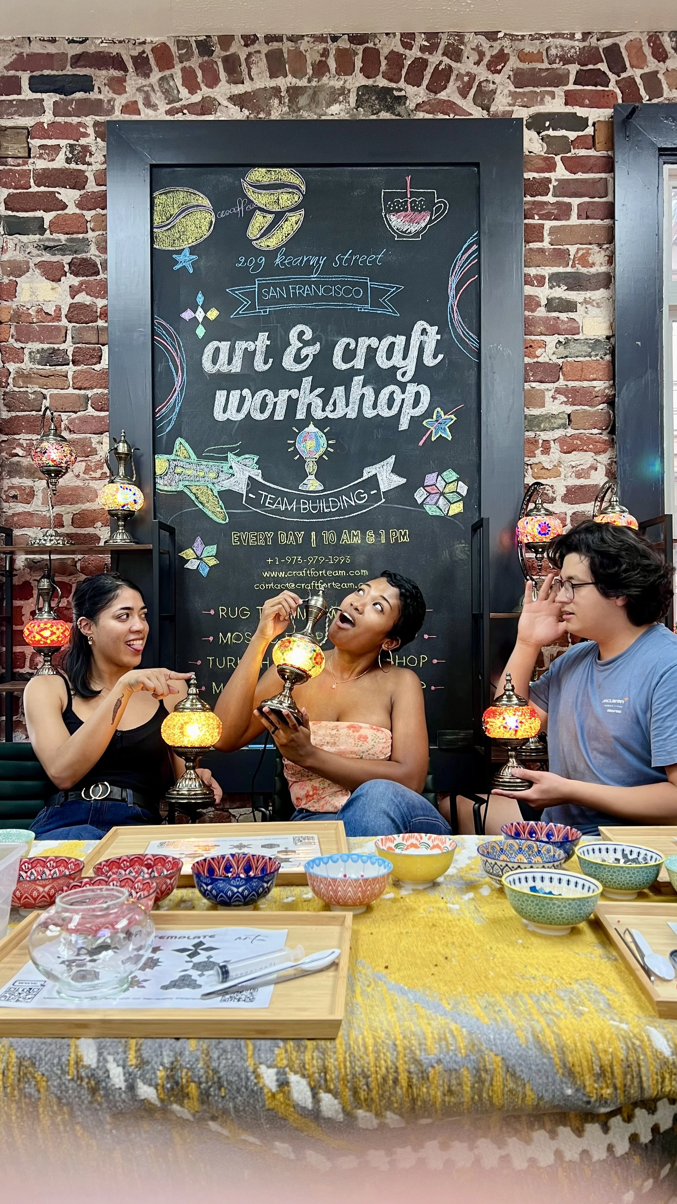 Three women sitting at a table during an art and craft workshop, holding colorful lamps, with a brick wall and a chalkboard sign behind them that reads 'art & craft workshop' in San Francisco.