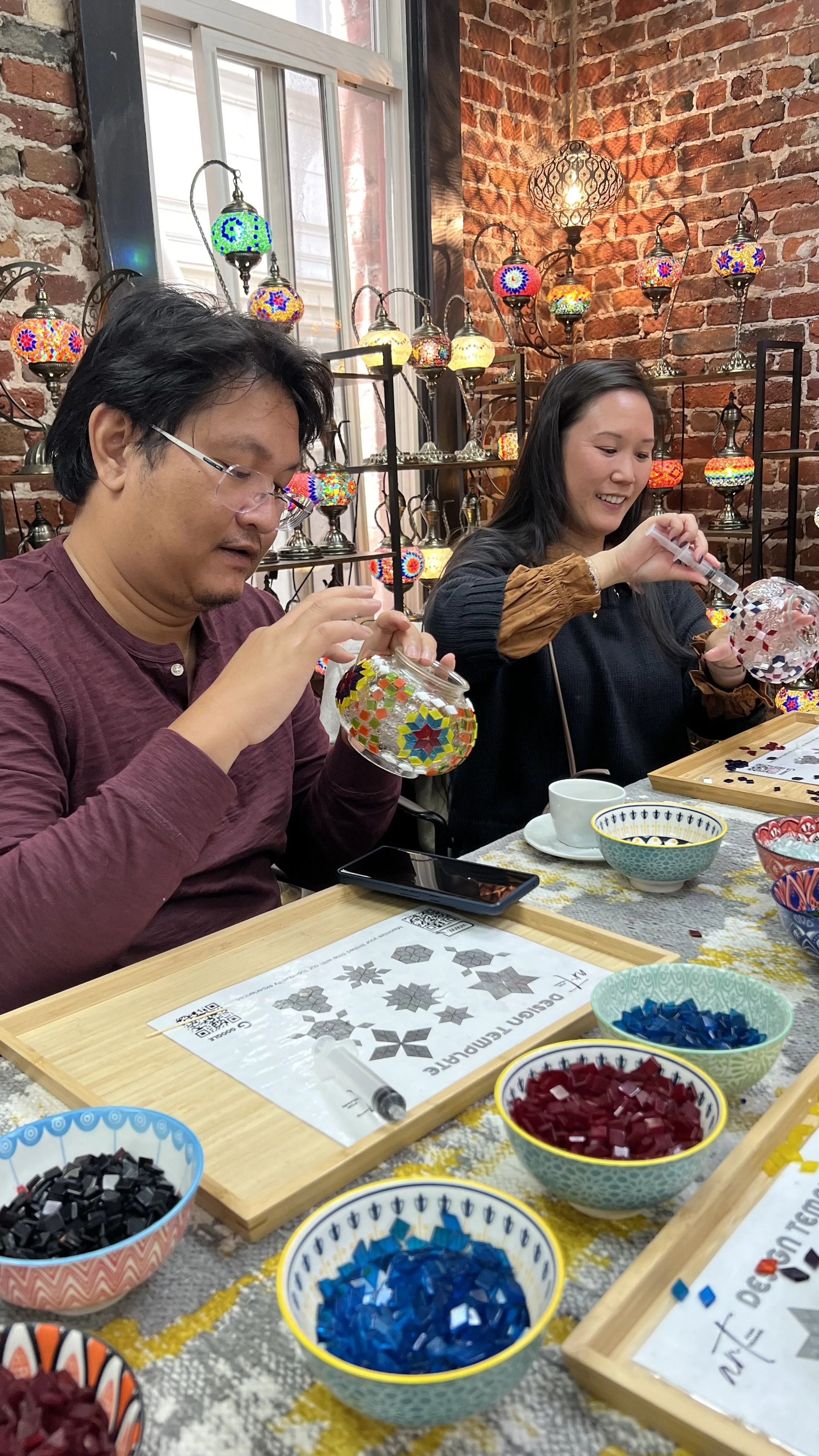 Two people sitting at a table, making colorful mosaic glass art with small pieces, surrounded by bowls of colored glass mosaics, in a cozy brick-walled shop decorated with colorful mosaic glass lamps.