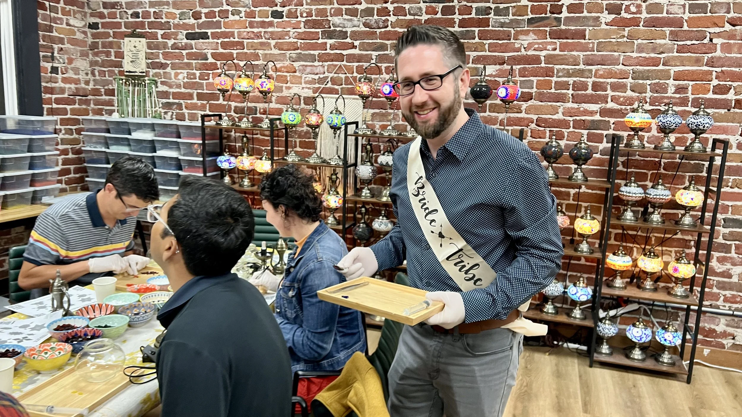 A man wearing glasses, a dark patterned shirt, and a sash that reads 'Bridal Trail' smiles while holding a tray, at a craft or jewelry-making event, with other people working at a table and decorative lamps on shelves behind him.