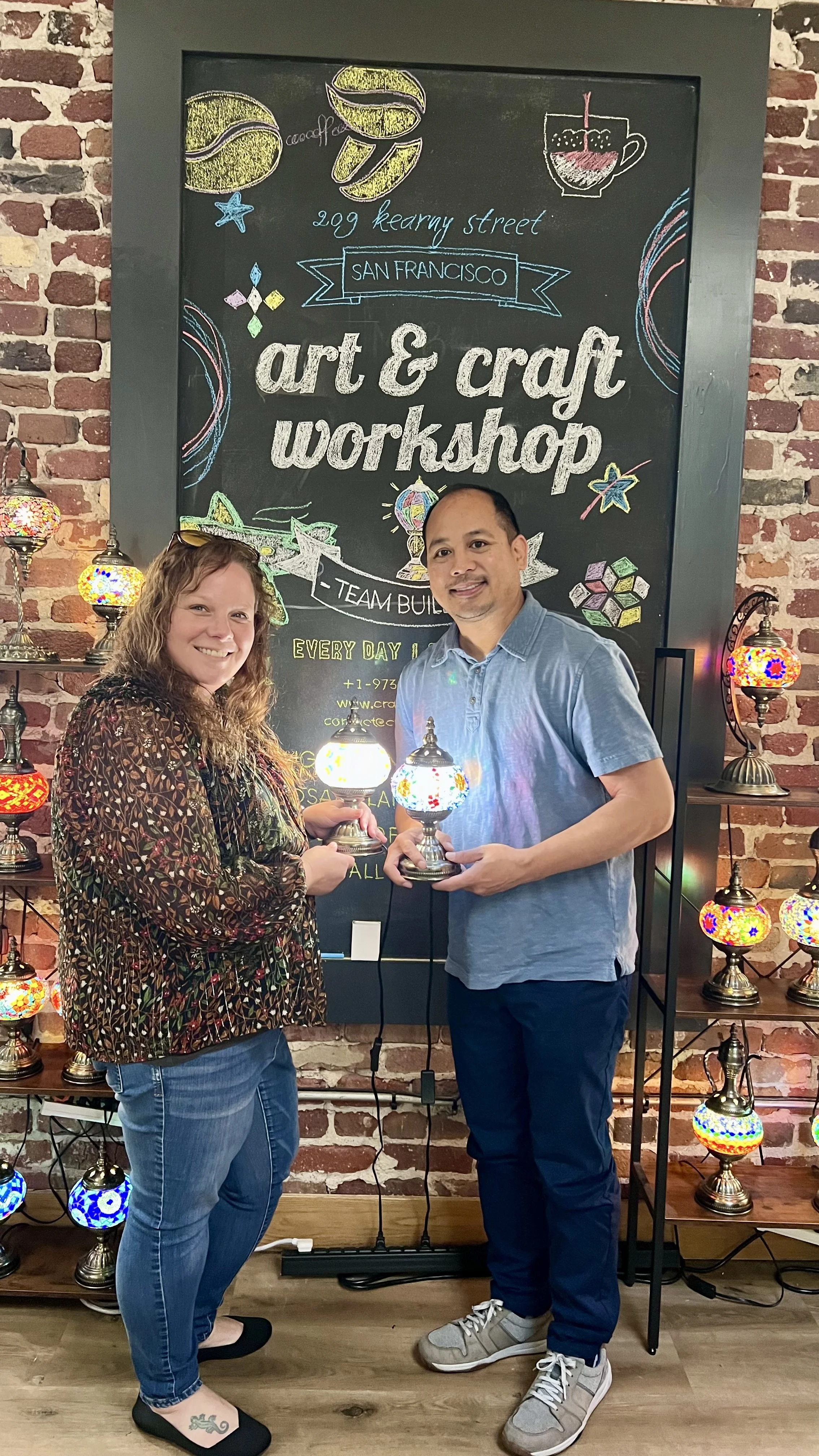 Two people smiling and holding glass lamps in front of a decorative chalkboard promoting an art and craft workshop, with shelves of colorful lamps and a brick wall in the background.