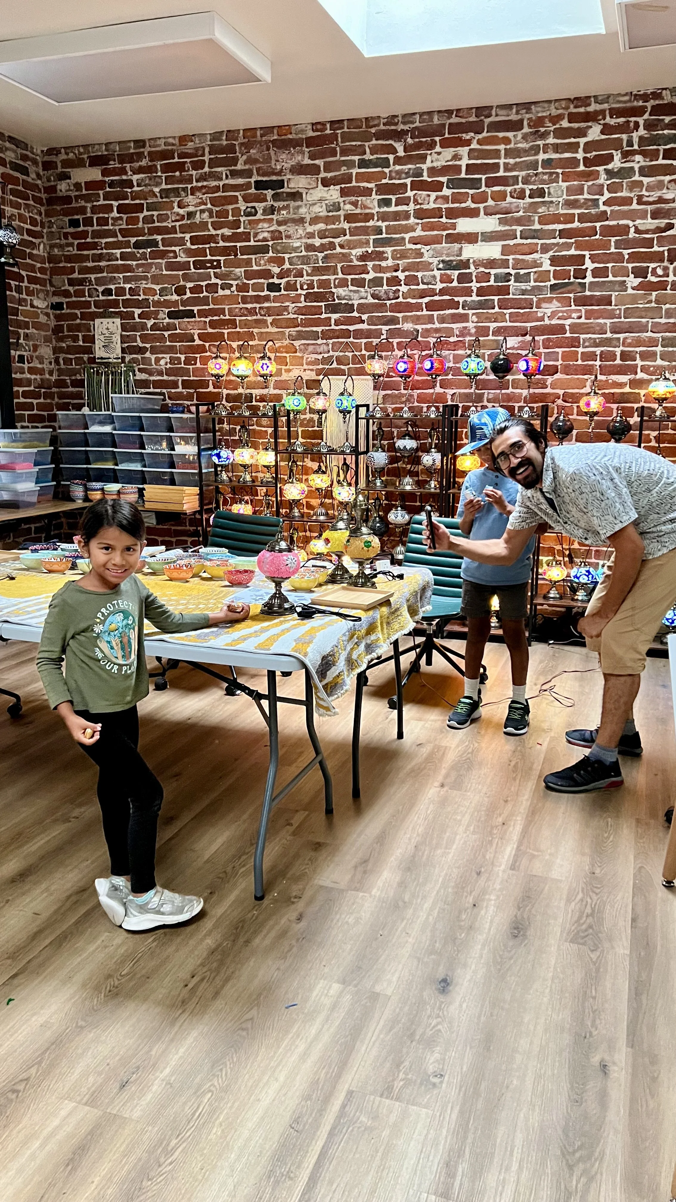 A young girl and two men are in a room with a brick wall and a display of colorful hanging glass lamps. The girl is smiling and standing by a table with lamps and bowls, one of the men is holding a phone and smiling, and the other man is standing in 
