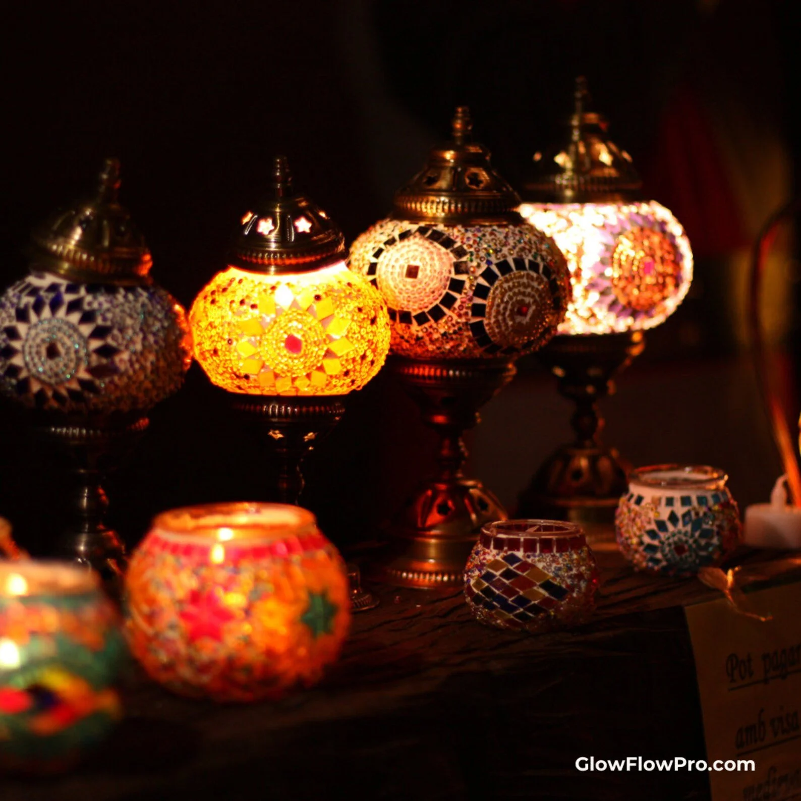 Colorful mosaic Turkish lamps glowing in a dark setting, arranged on a table, with some smaller mosaic glass candle holders in front.