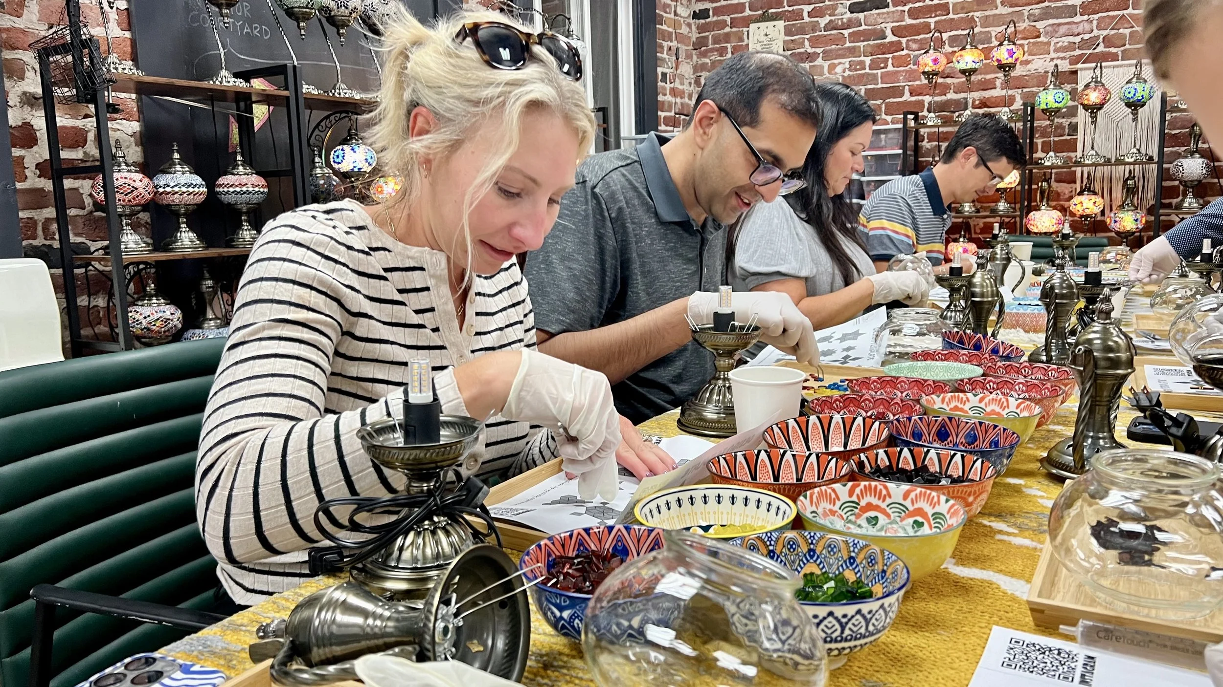 Group of people participating in a lamp decorating workshop at a table with colorful decorated bowls and lamps, in a brick-walled craft studio.