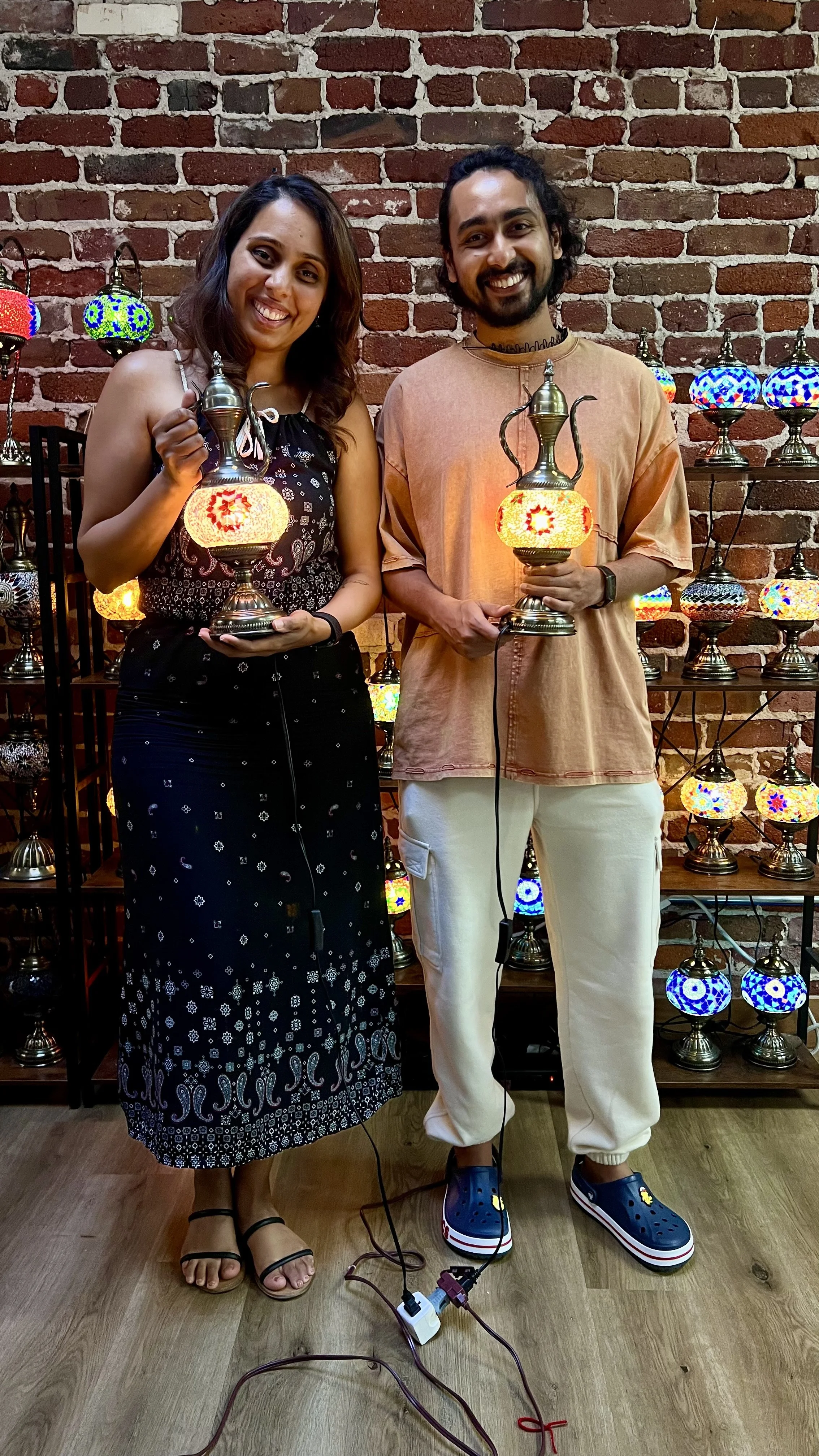 Two smiling people holding decorative lamps, standing in front of a brick wall with illuminated mosaic lamps on shelves behind them.