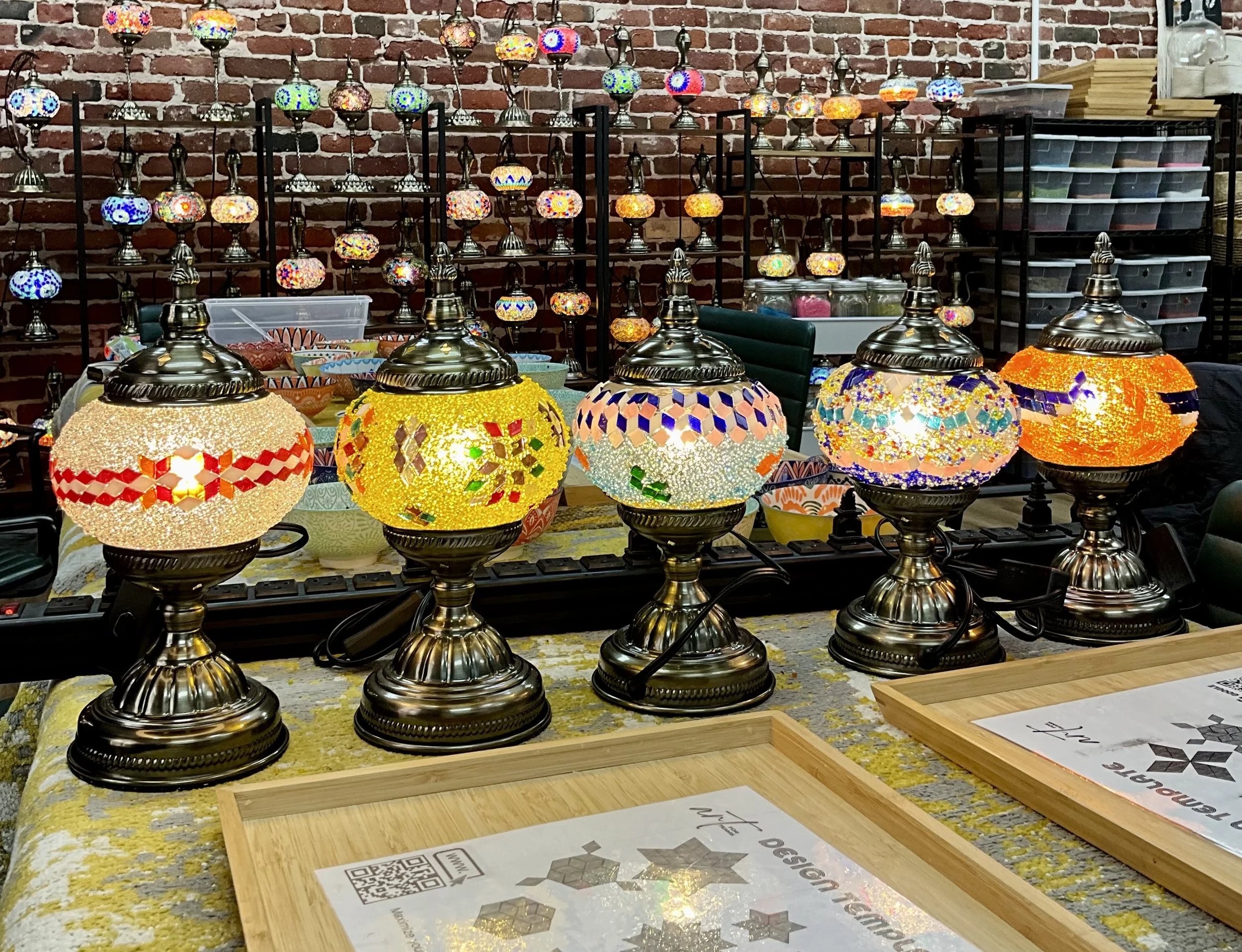 Colorful mosaic glass table lamps on a craft table, with additional mosaic lamps displayed on shelves in the background. The lamps feature various vibrant patterns and colors, and the setting appears to be a craft or decor shop.