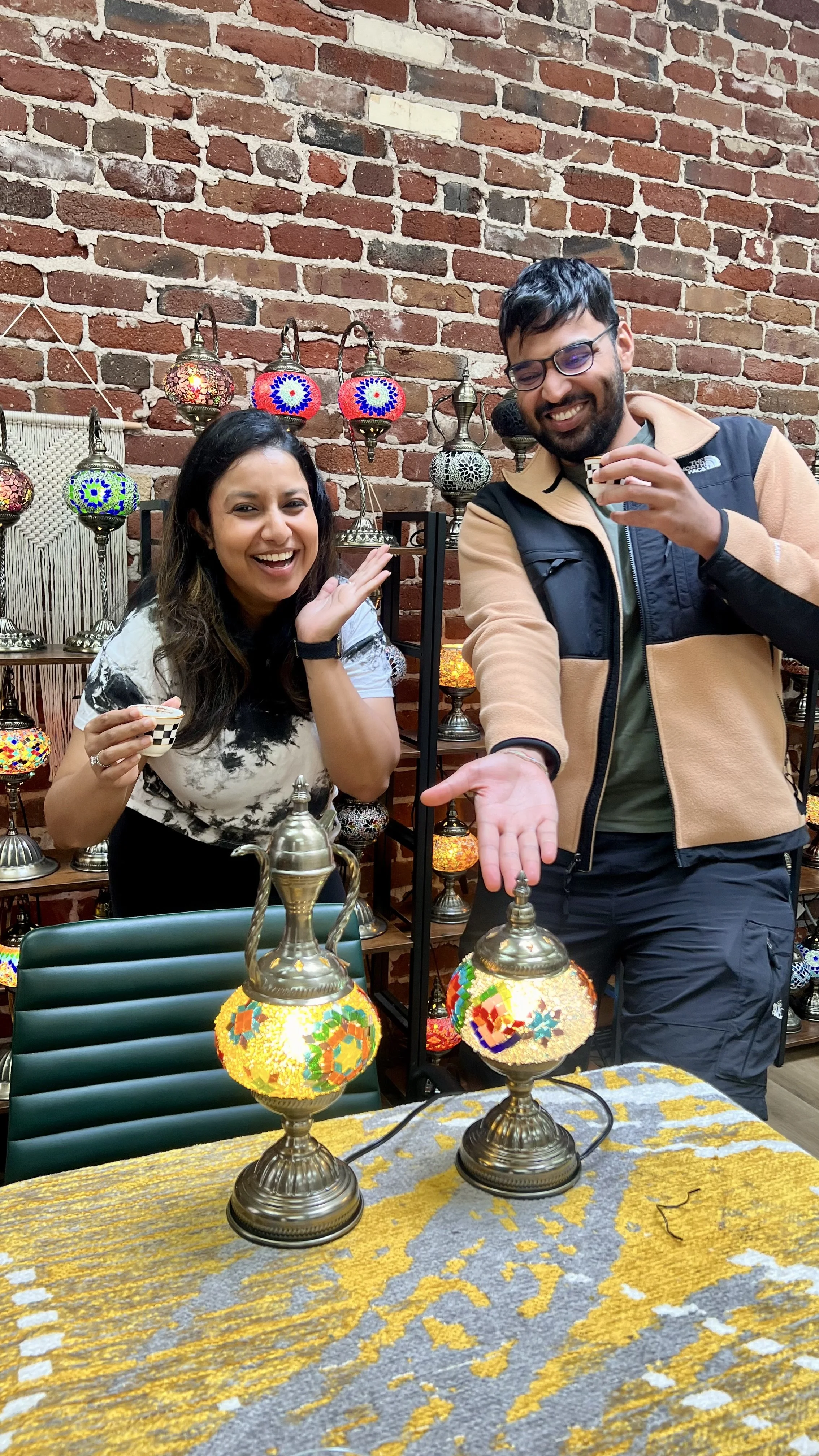 Two smiling people, a woman with long dark hair and a man with glasses, posing with decorative mosaic lamps, in front of a brick wall, with a table and colorful lanterns in the background.