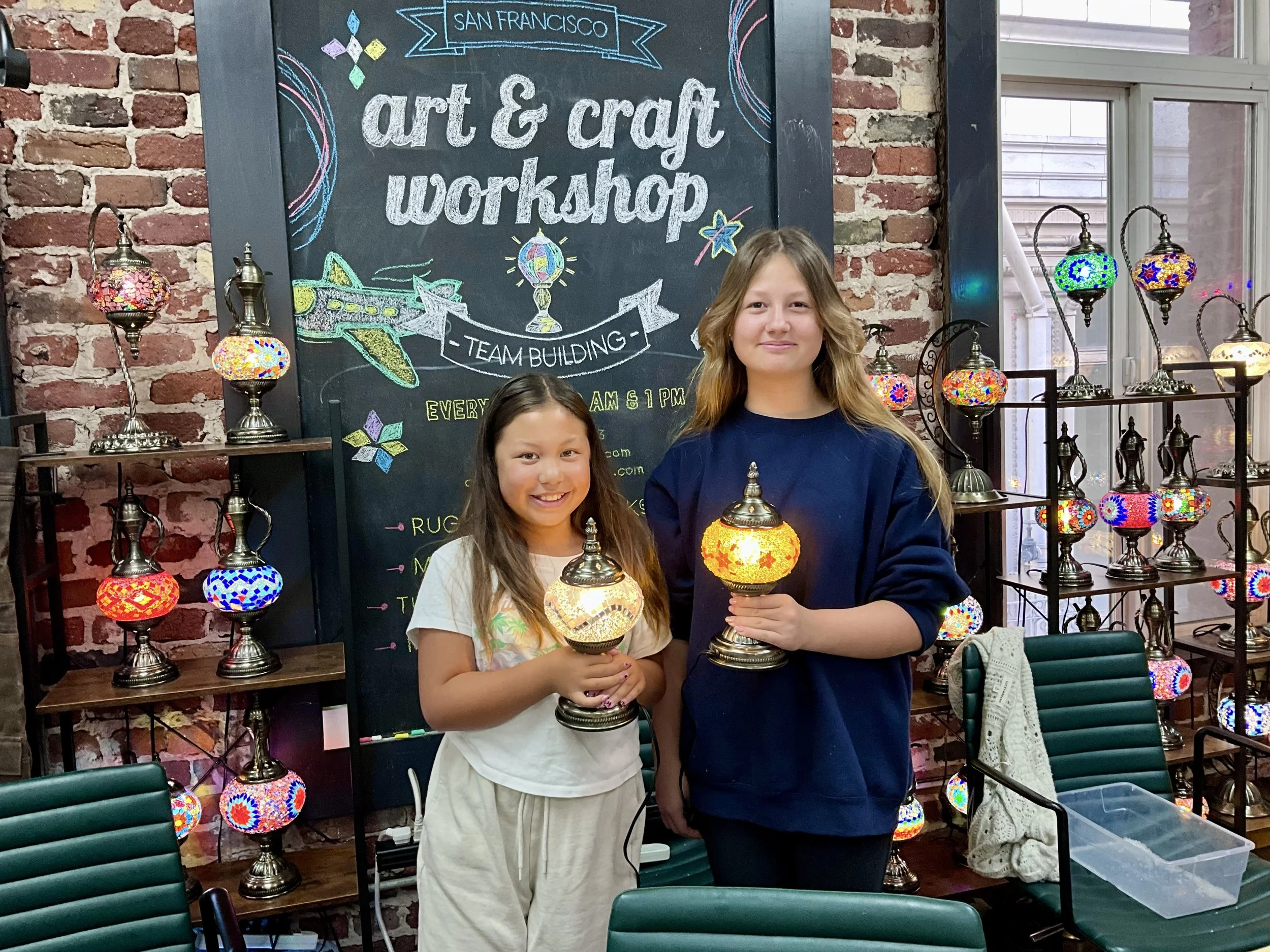 Two girls holding colorful mosaic lamps at an art and craft workshop in front of a blackboard sign that reads 'San Francisco Art & Craft Workshop'. The workshop is set against a red brick wall with shelves of colorful mosaic lamps.
