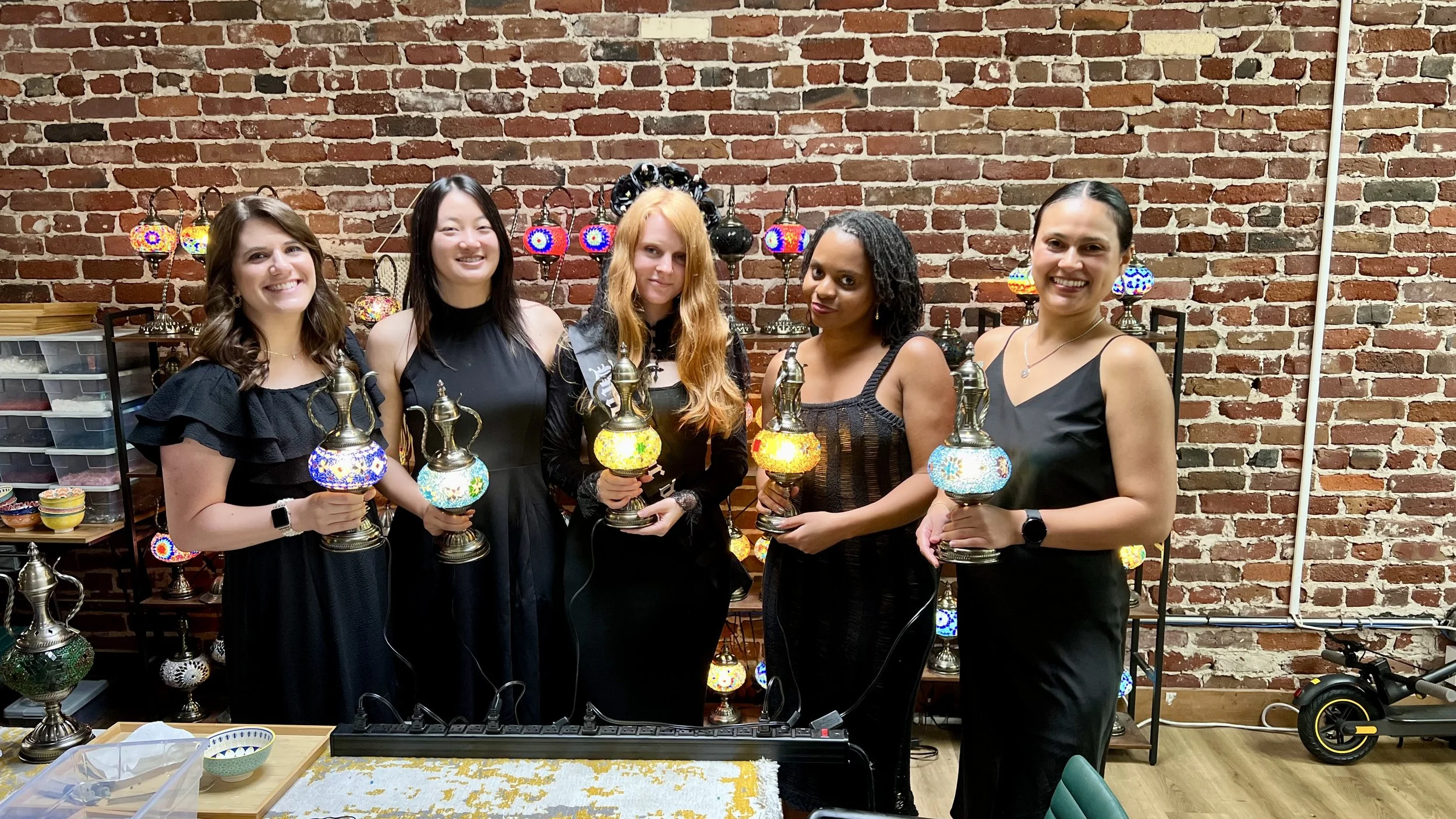 Five women standing in front of a brick wall holding colorful Turkish mosaic lamps as trophies, smiling at the camera.
