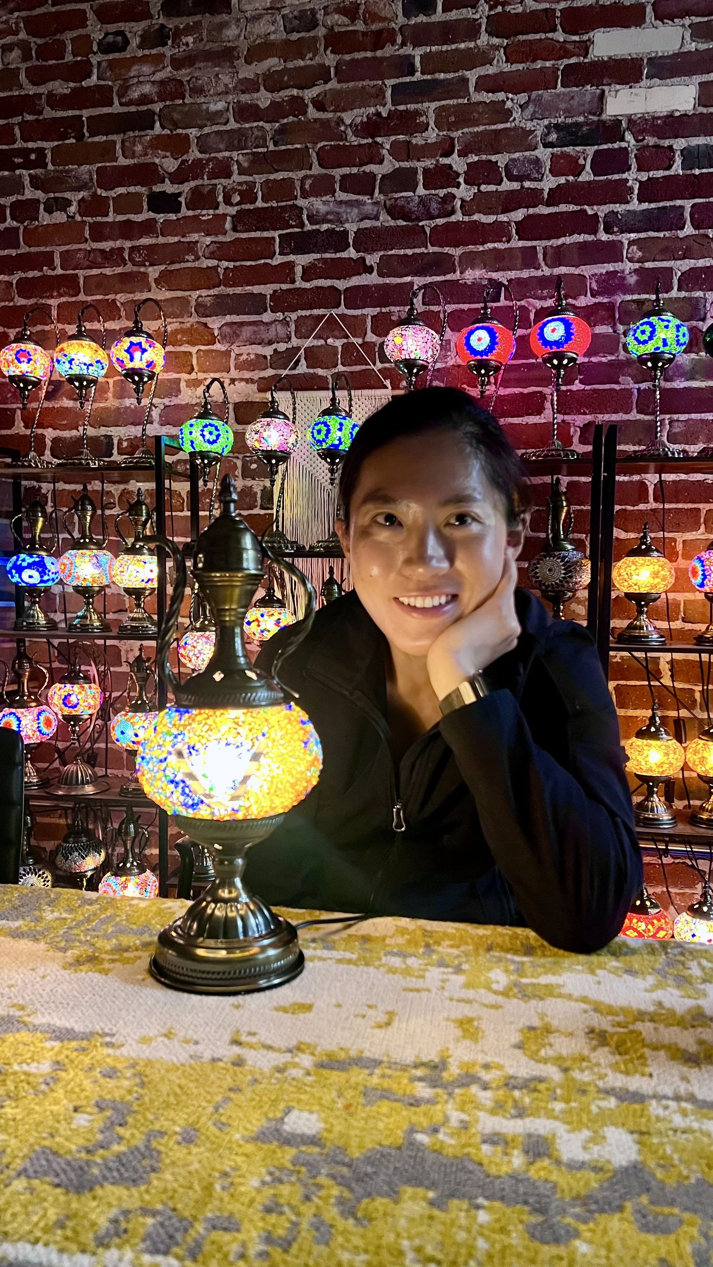 Smiling woman sitting at table with colorful mosaic Turkish lamps in the background.