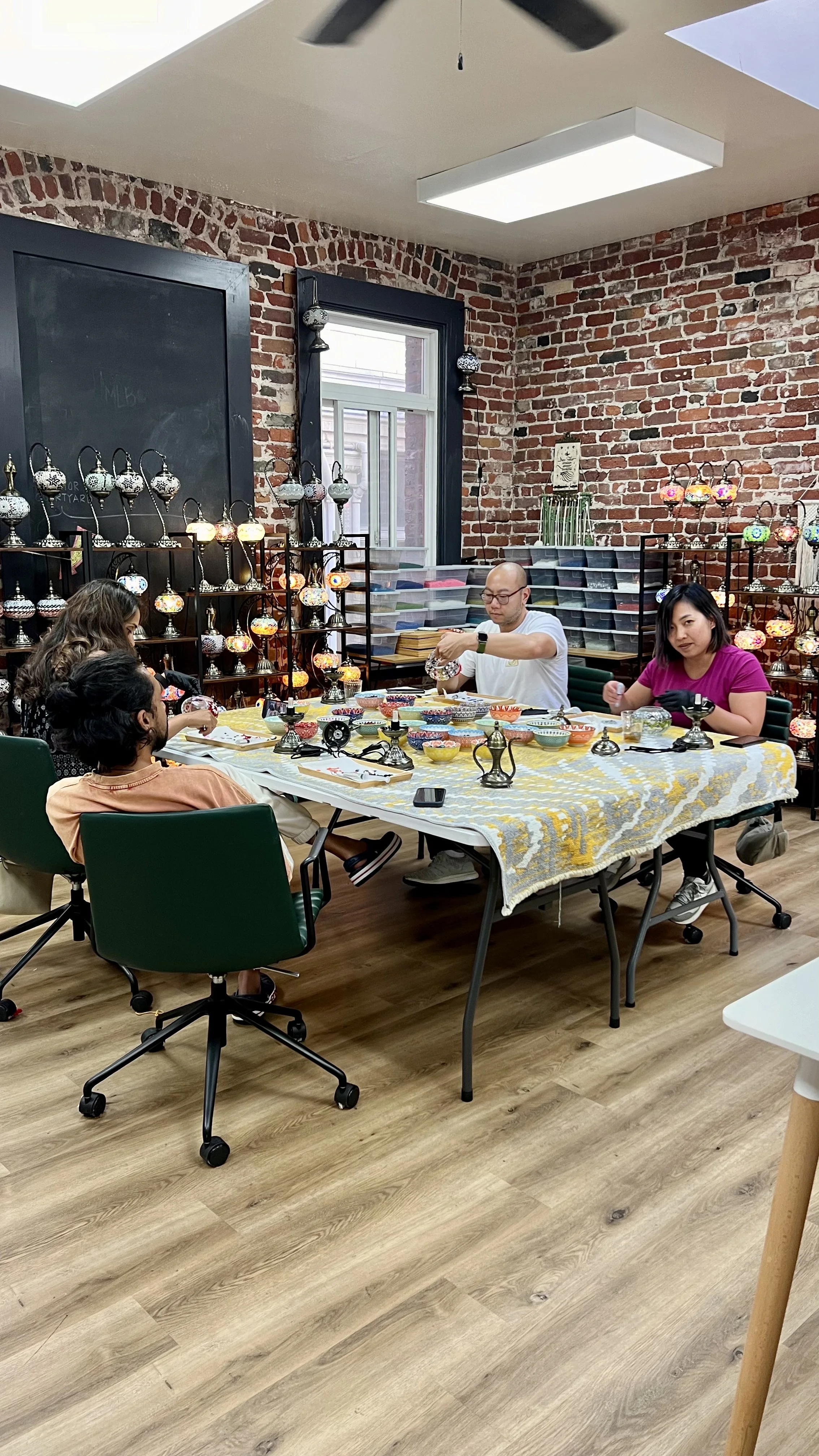 People painting glass lampshades on a table in a workshop with brick walls and colorful lamps displayed on shelves