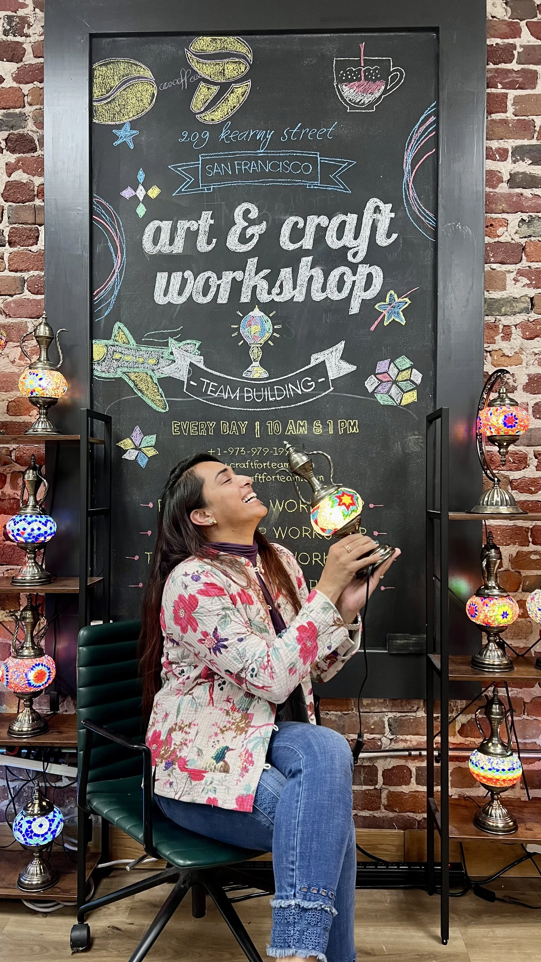 Woman laughing and holding a decorative lamp in art and craft workshop with a blackboard sign about team building. The workshop is held at 209 Kearny Street, San Francisco.