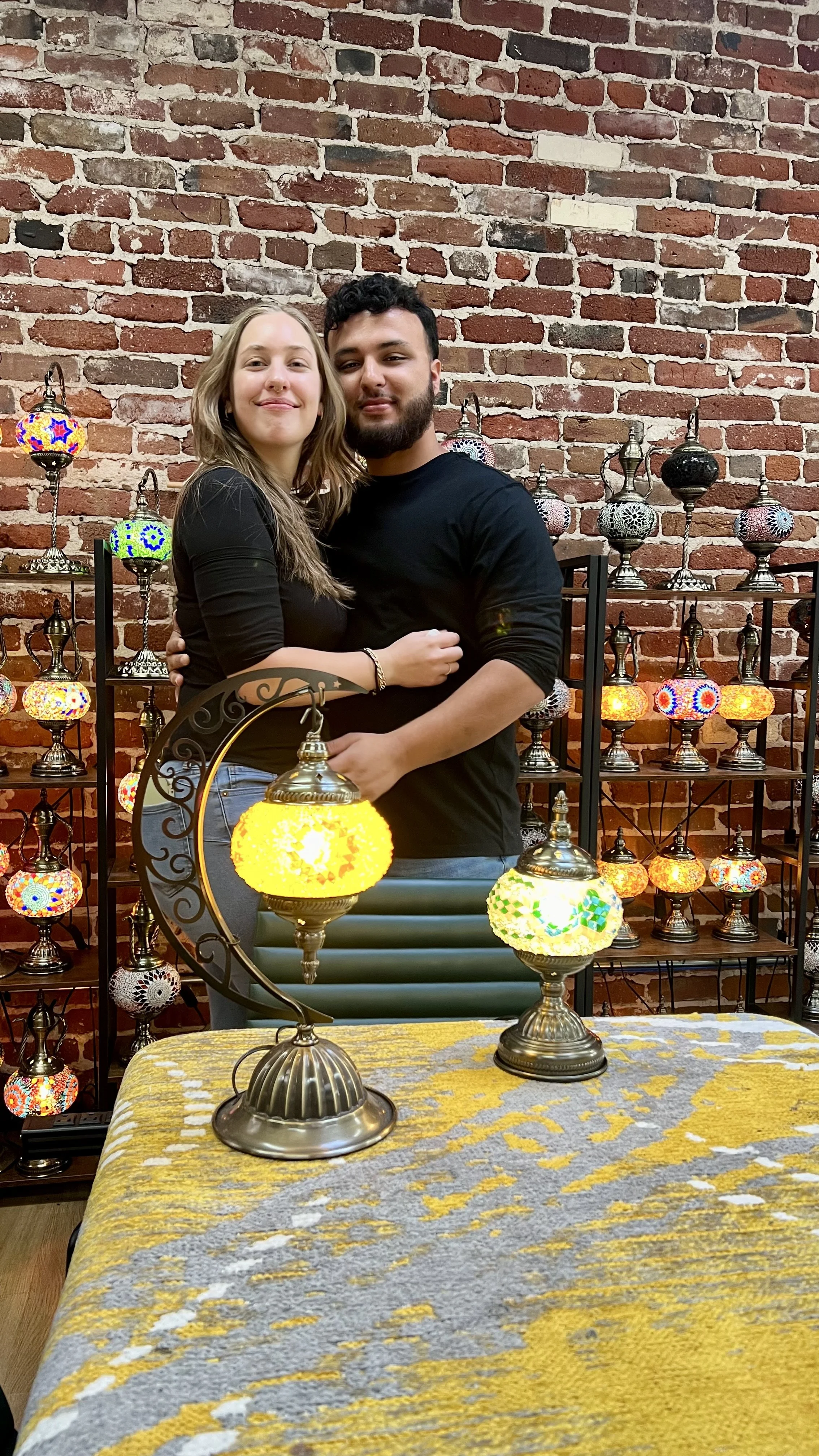 A young couple is standing close together, smiling, with a display of colorful Turkish mosaic lamps and a brick wall in the background. The table in front has decorative Turkish lamps.