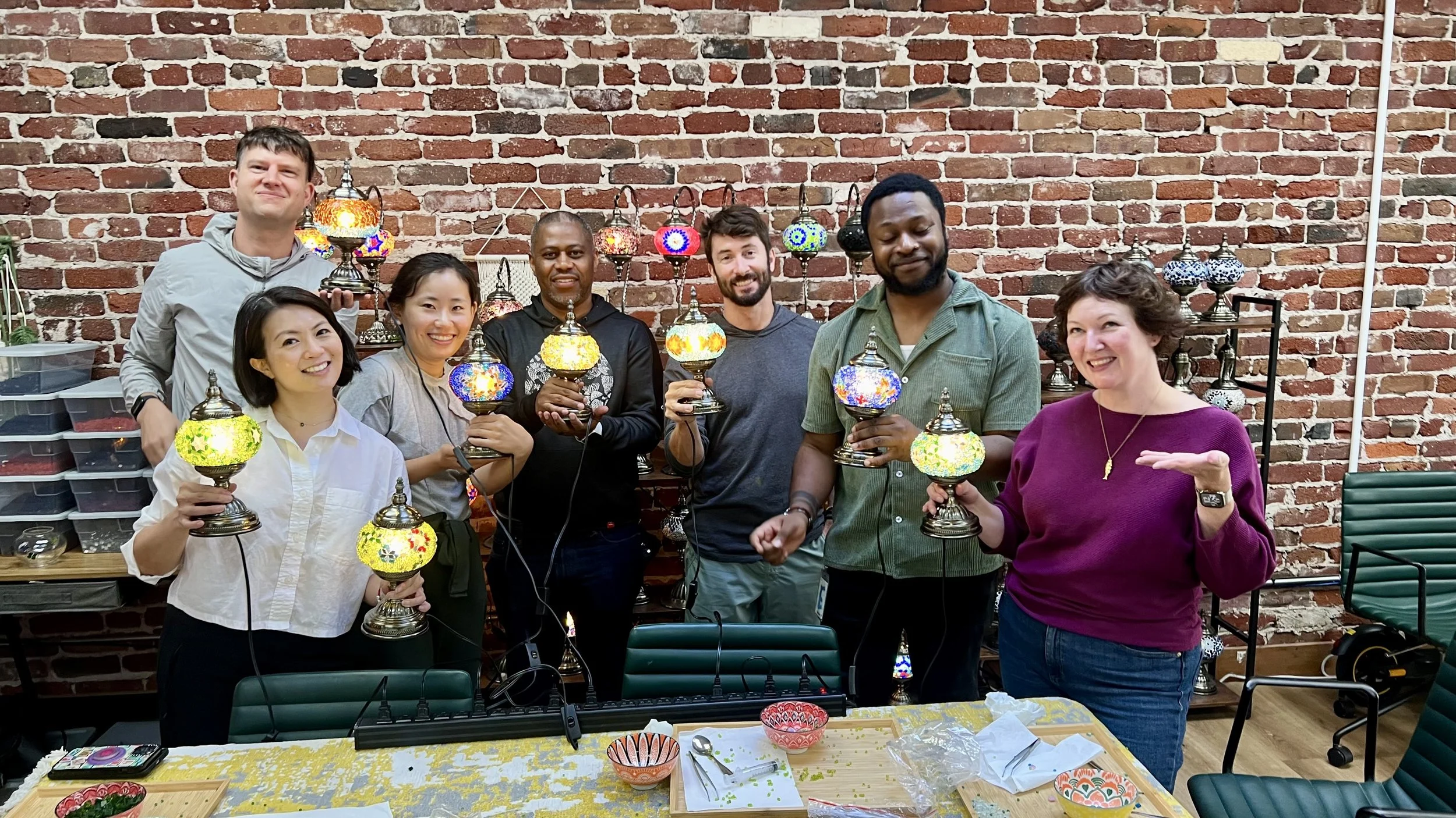 A diverse group of eight people posing together indoors, holding colorful mosaic lamps, with a brick wall and display of lamps in the background.