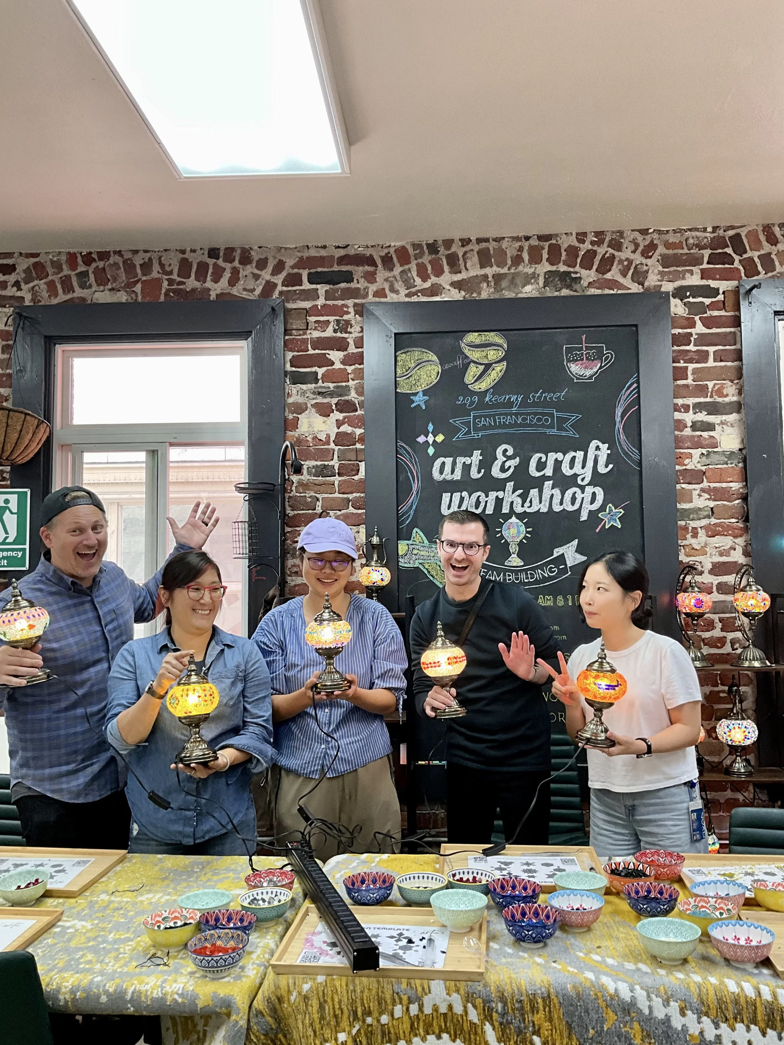 Group of six people smiling and holding colorful mosaic lamps in a craft workshop with a blackboard that reads 'art & craft workshop' behind them.
