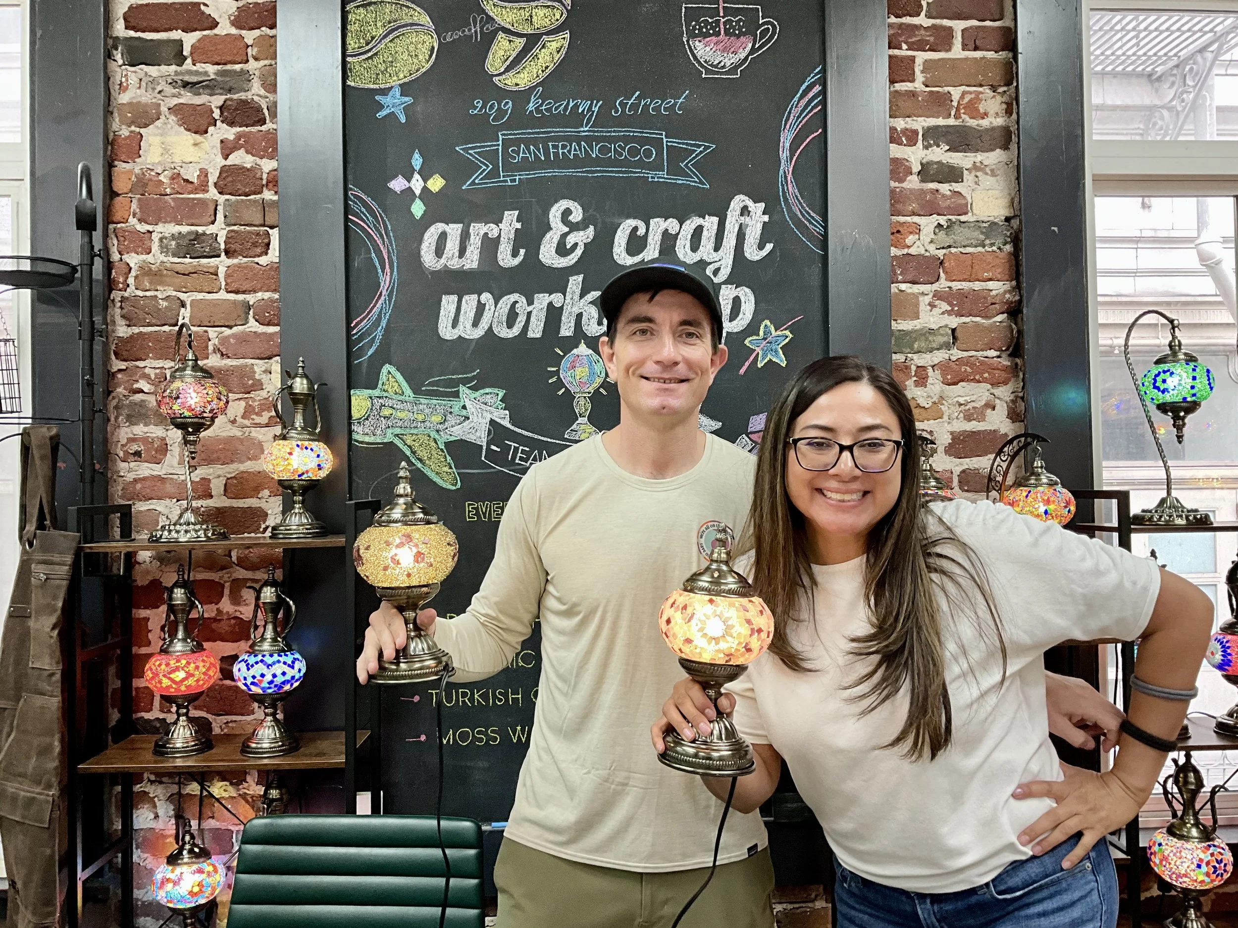 Two smiling people, a man and a woman, holding colorful mosaic lamps in front of a chalkboard with decorative writing about an art and craft workshop in San Francisco. The background features a brick wall and additional mosaic lamps.