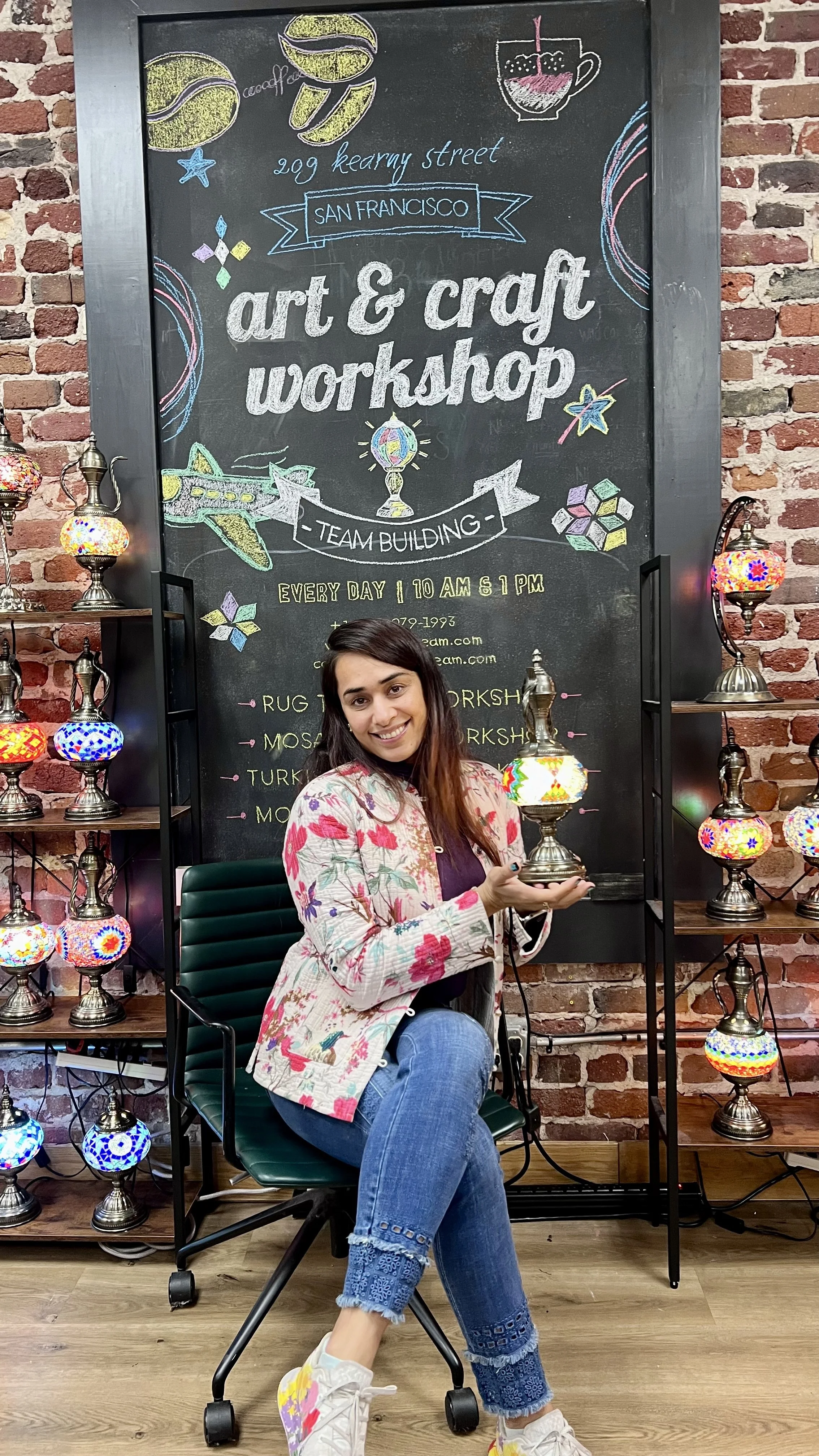 A smiling woman holding a colorful lamp sits in front of a blackboard advertising an art and craft workshop. The workshop is held every day from 10 am to 1 pm on Kearny Street in San Francisco. The blackboard features colorful chalk drawings of lamps