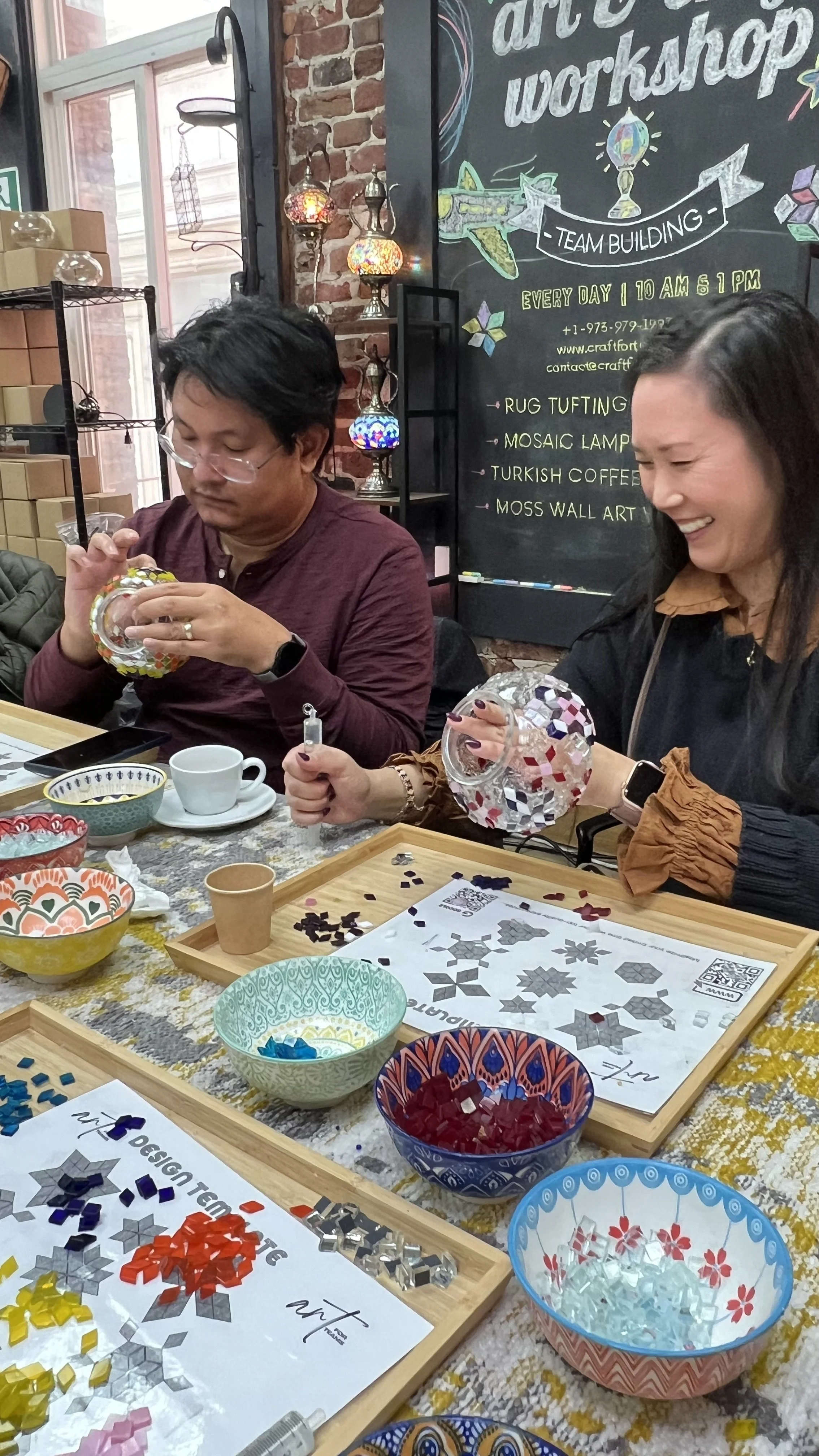 Two people are engaging in a mosaic glass crafting workshop at a table filled with colorful mosaic tiles, bowls, and paper templates. They are handling glass pieces while smiling and focused on their craft, inside a room with a brick wall and decorat
