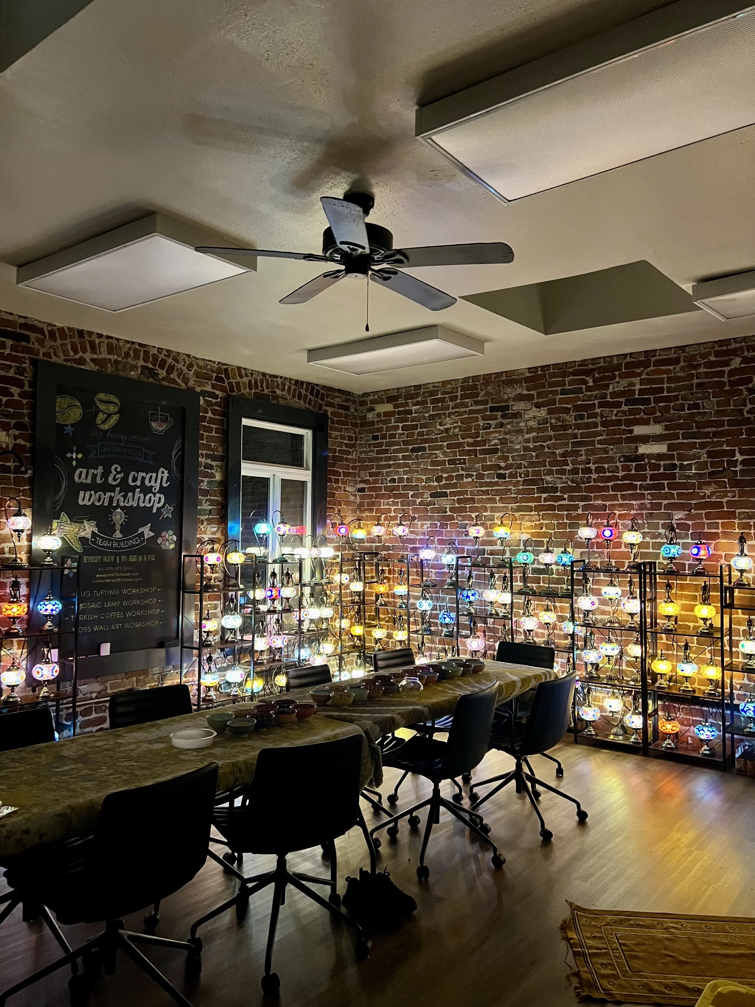 A room with brick walls has colorful LED lamps on black shelves arranged in a semi-circle. A table with black chairs surrounds it, and a chalkboard sign on the wall advertises art and craft workshops.