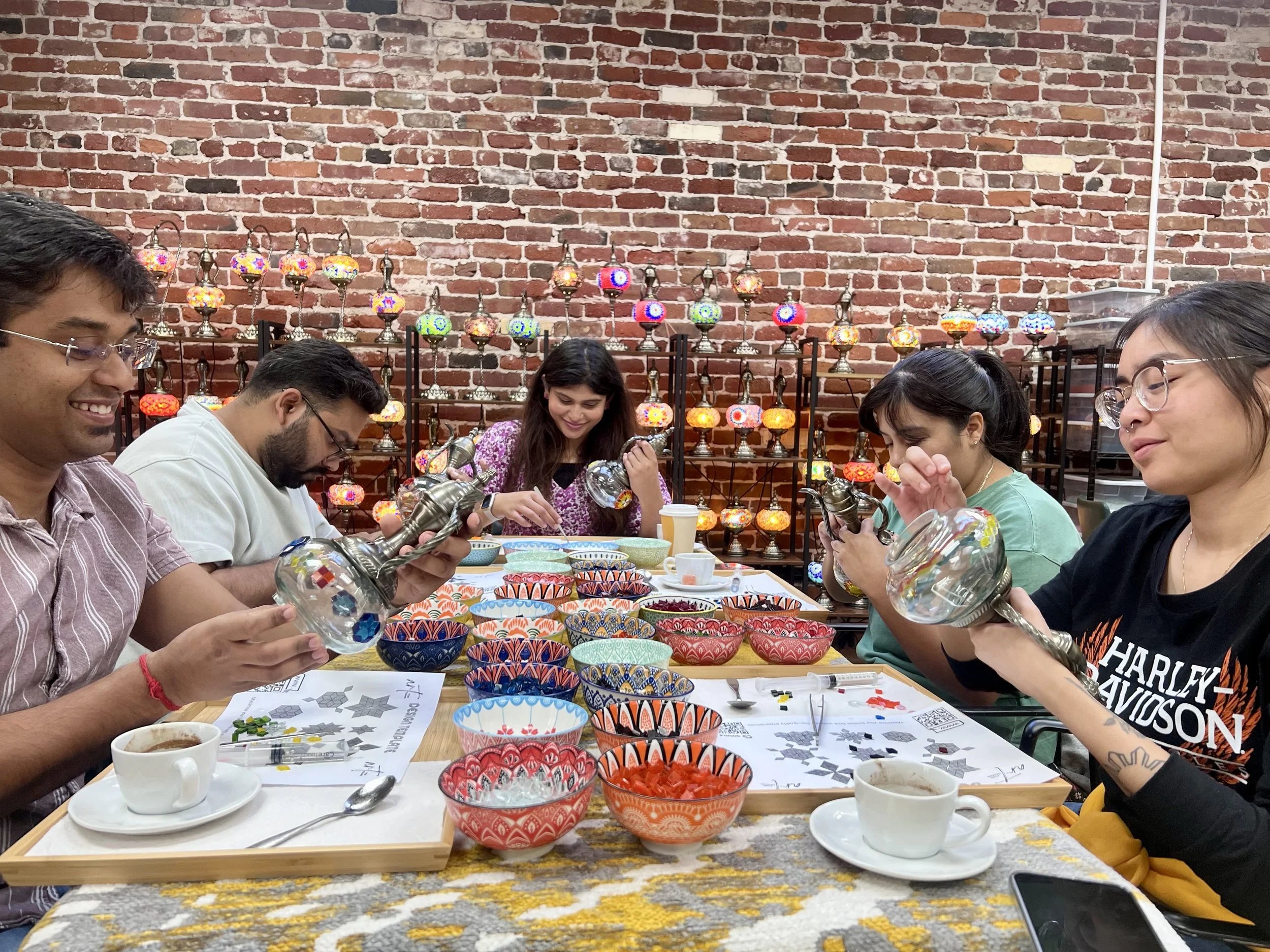 Group of five people sitting at a table with colorful handcrafted bowls and decorations, engaged in decorating glass lanterns, in a room with a brick wall background decorated with lit mosaic lamps.