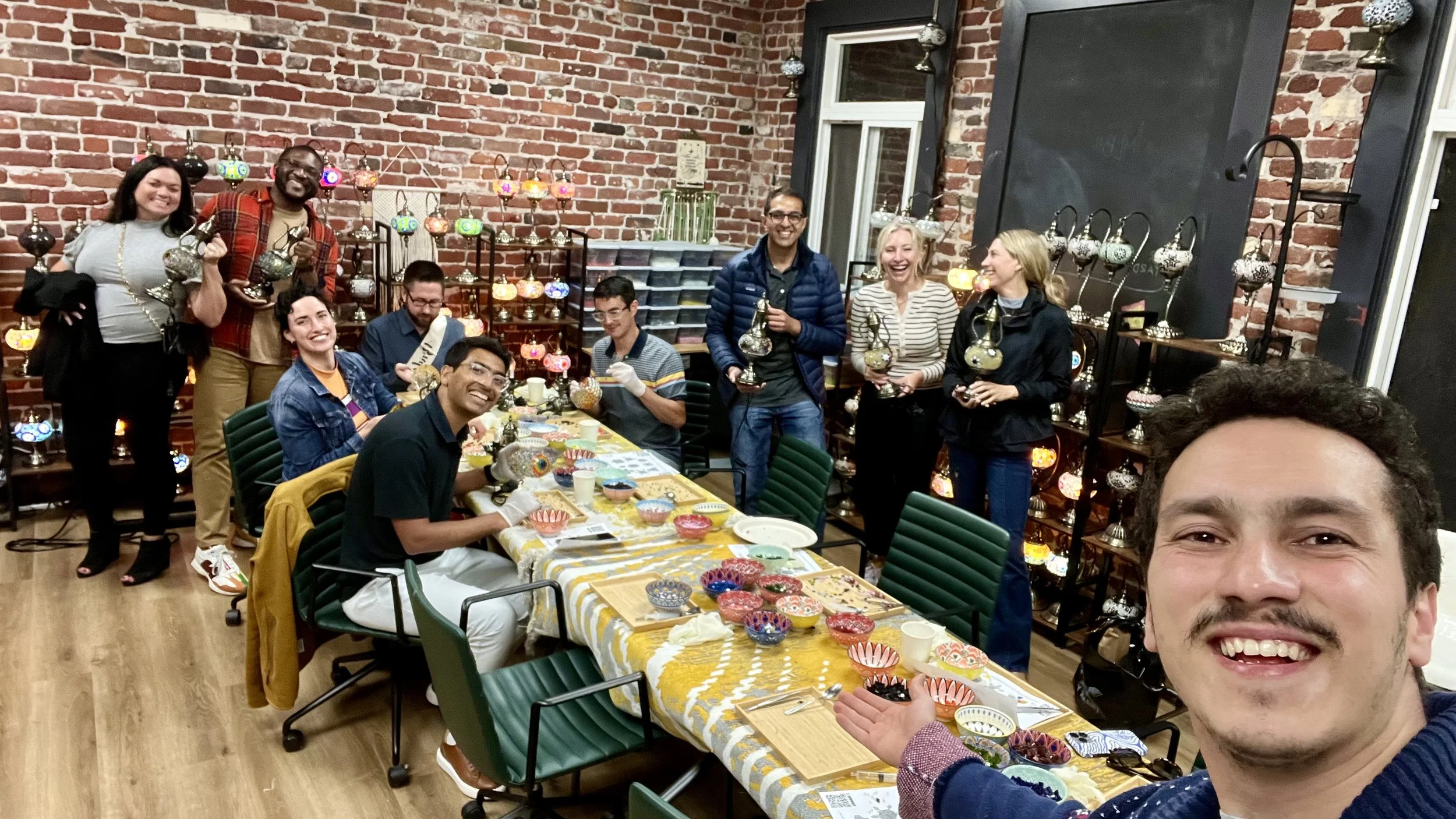 Group of people gathered around a table with decorated ceramic bowls, in a room with exposed brick walls and shelves of colorful lamps, celebrating an arts and crafts activity.
