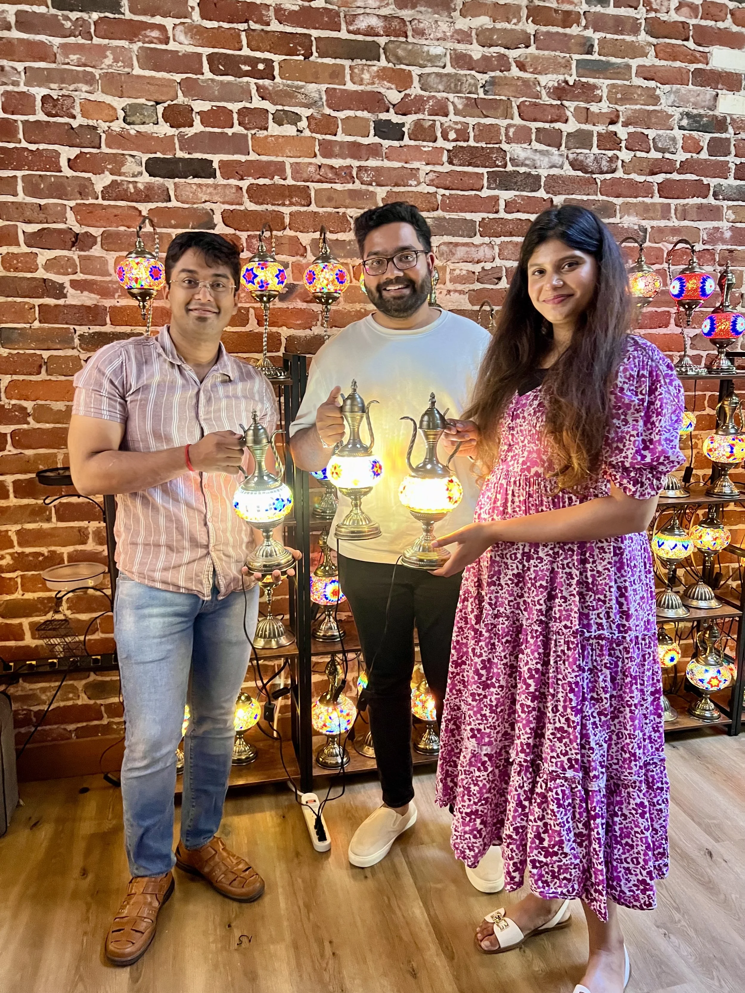 Three people holding decorative lamps indoors against a brick wall with lit colorful mosaic lamps on shelves behind them.