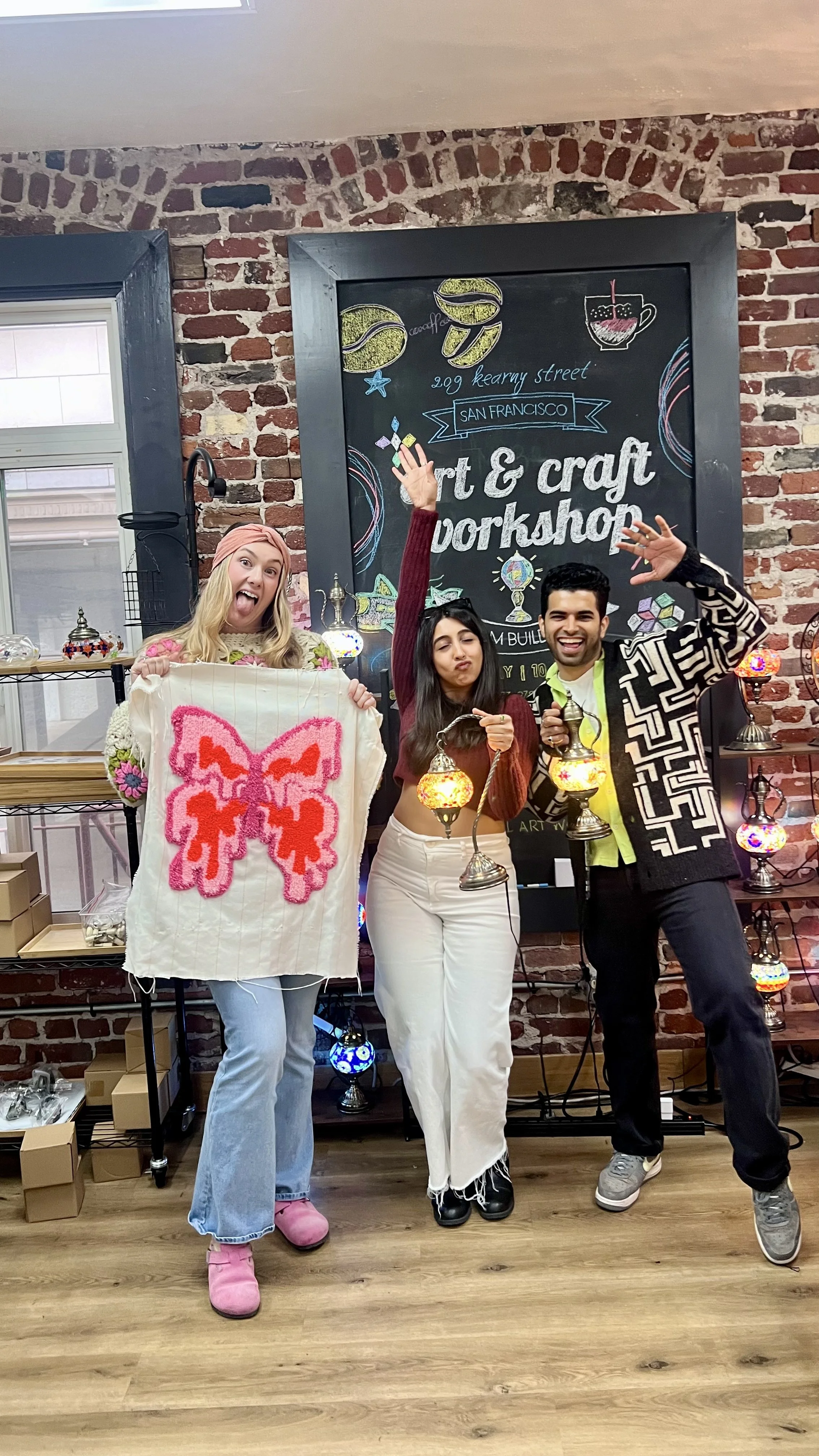 Three friends at an art and craft workshop holding decorative lanterns and a quilt with a butterfly design, smiling and waving, with a brick wall and a chalkboard sign behind them.