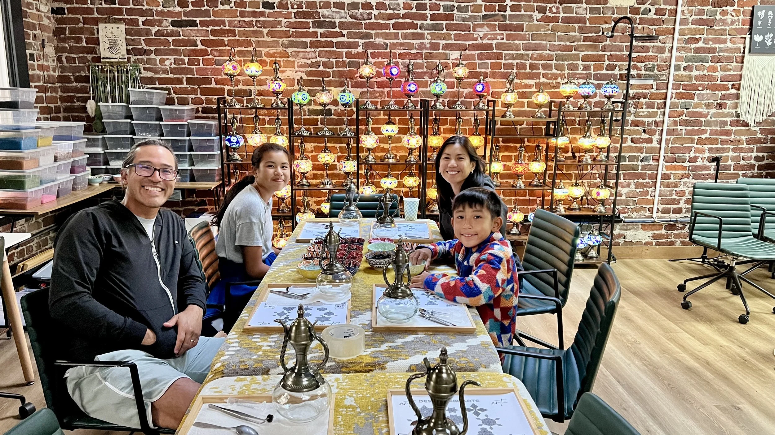 Four people sitting at a dining table surrounded by colorful Turkish lamps displayed on shelves against a brick wall.