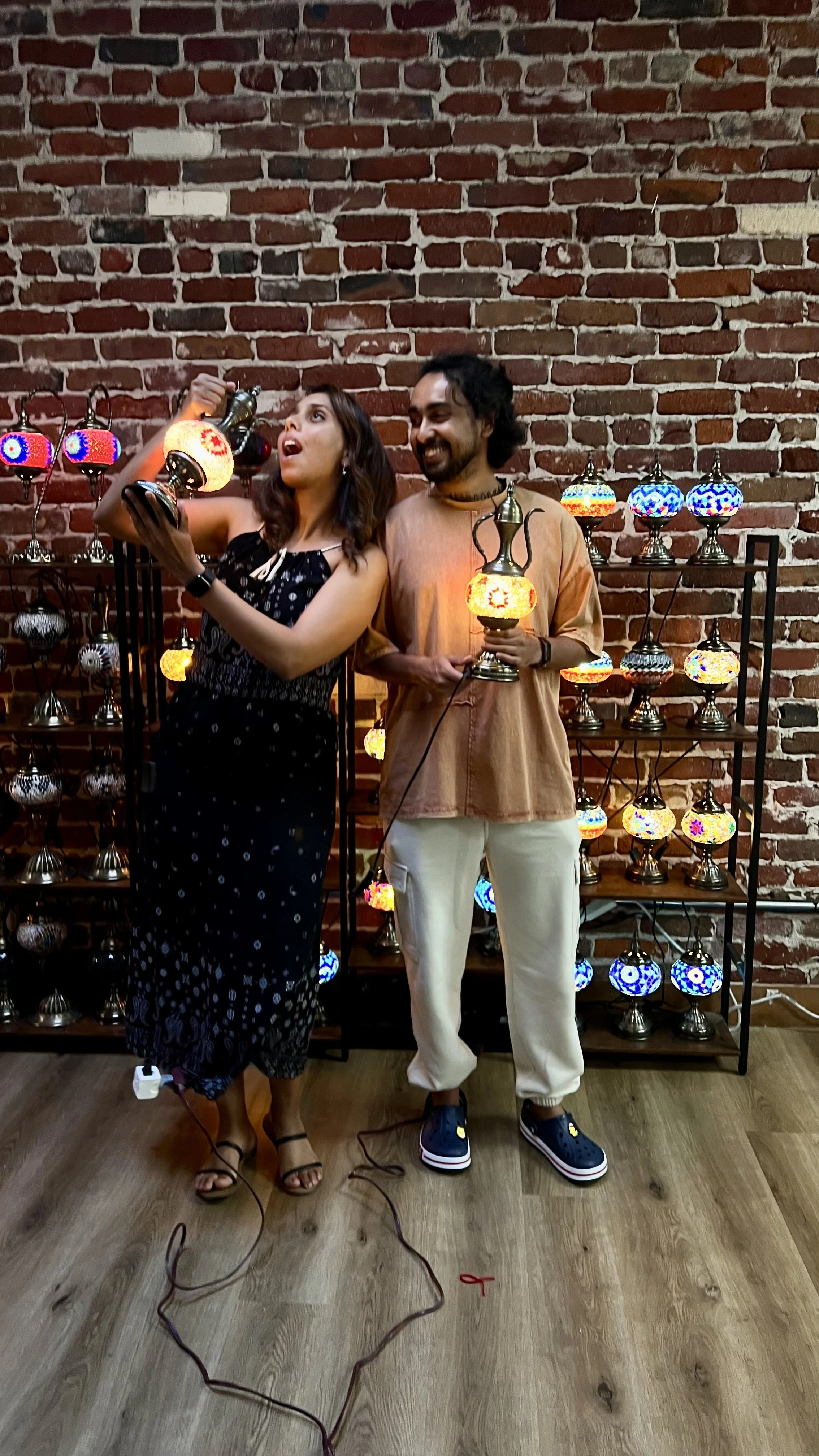 Two people, a woman and a man, are standing in front of a brick wall and a shelf of colorful lamps. The woman is holding a lamp and looking excited, while the man is holding a lamp and smiling.