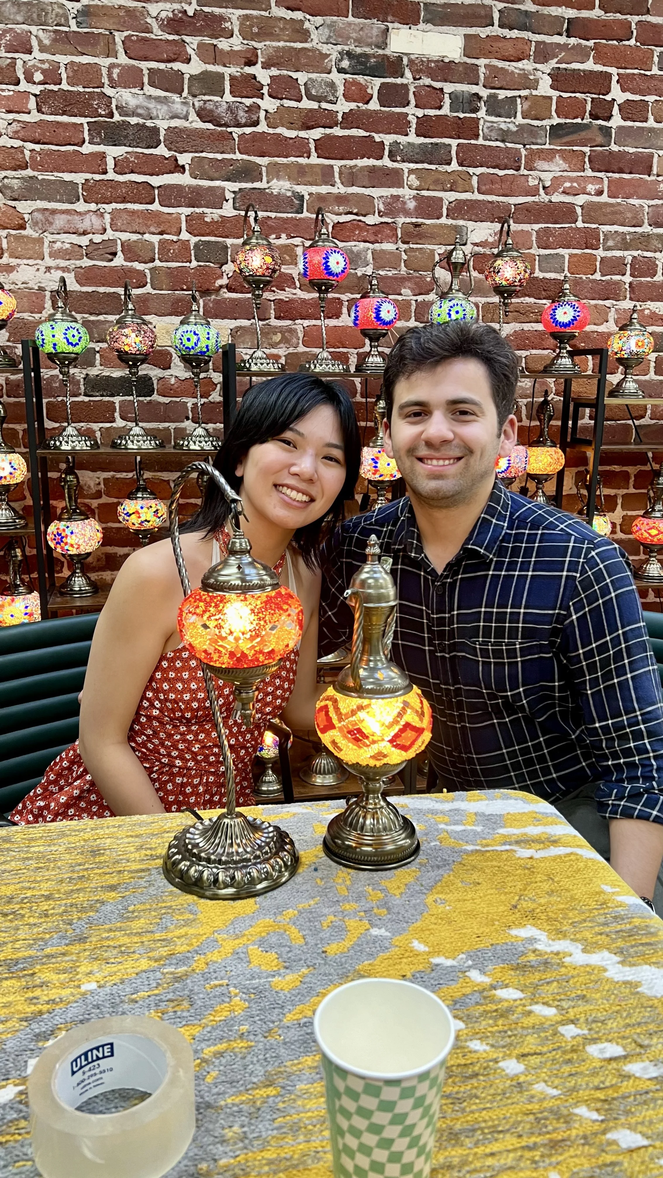 A smiling woman and man sitting at a table with decorative Turkish lamps and a brick wall background, with a cup and tape on the table.
