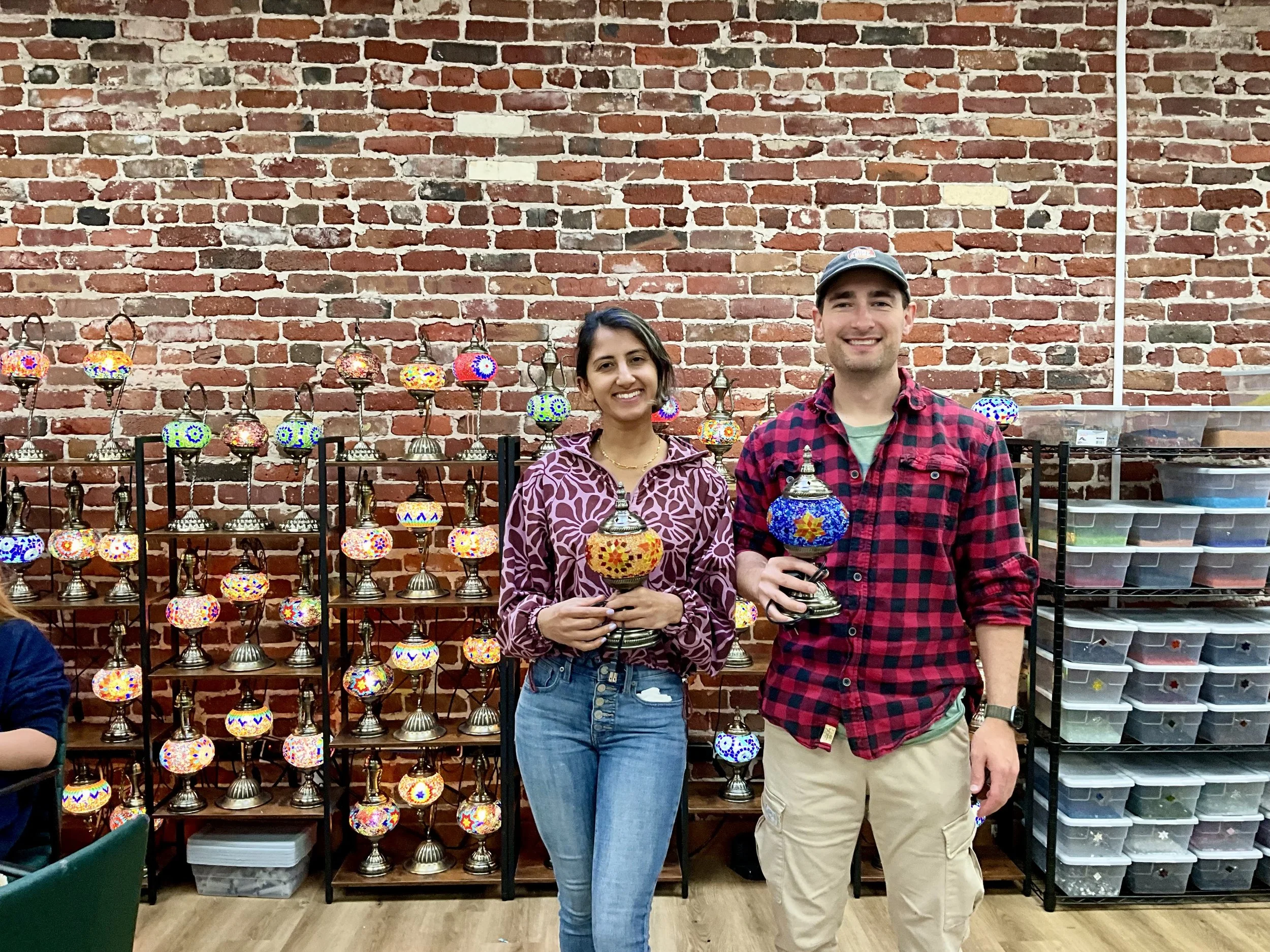 Two people standing in front of a brick wall with display shelves filled with colorful Turkish lamps. The woman on the left is smiling and wearing a purple patterned hoodie and jeans, holding a decorative lamp. The man on the right is smiling, wearin