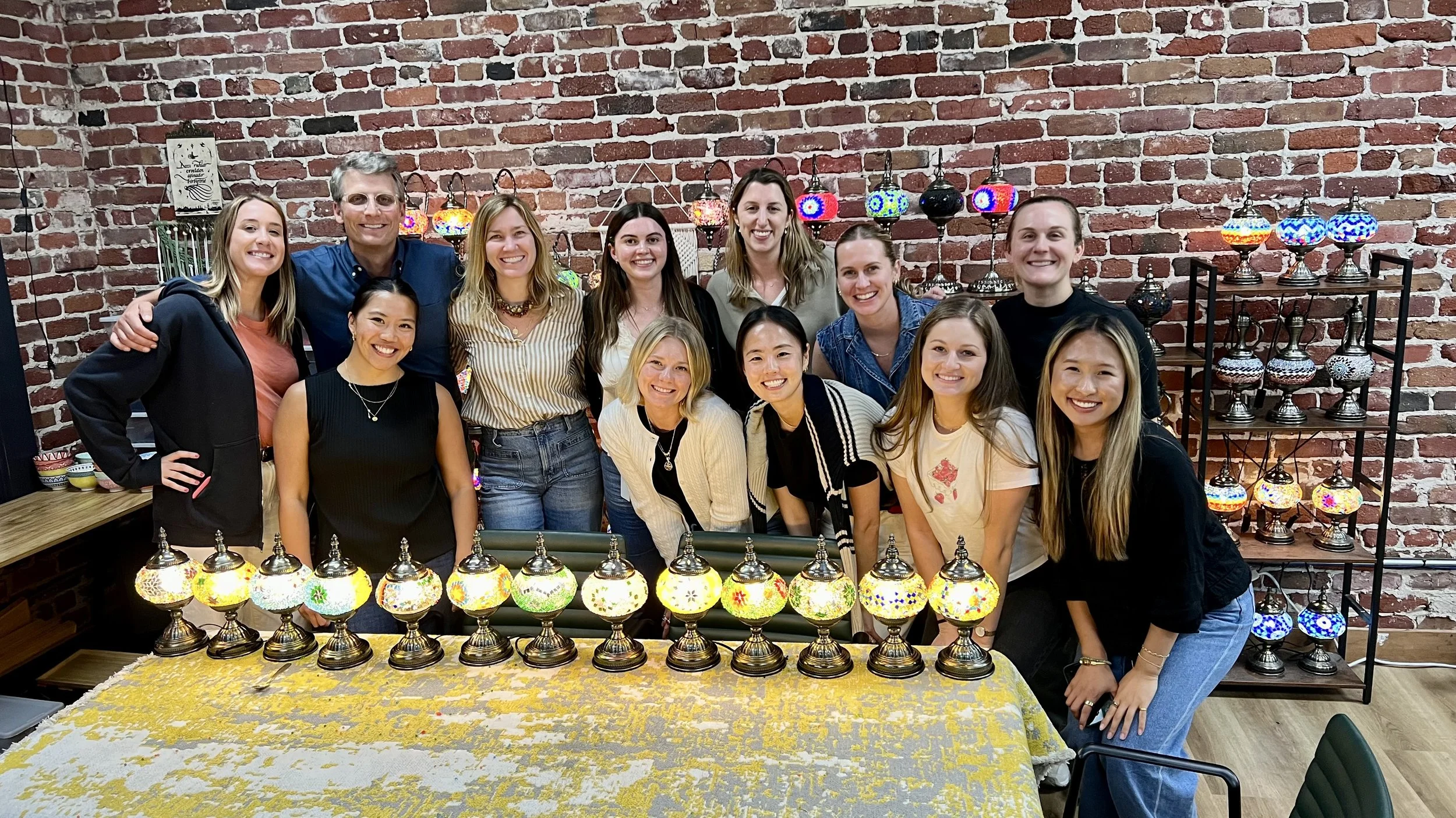 Group of 12 people smiling in front of a display of colorful glass lamps in a brick-walled room