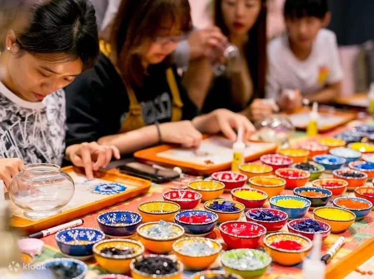 Children and adults making colorful painted mosaic lamp art at a craft table with bowls of paint and brushes.