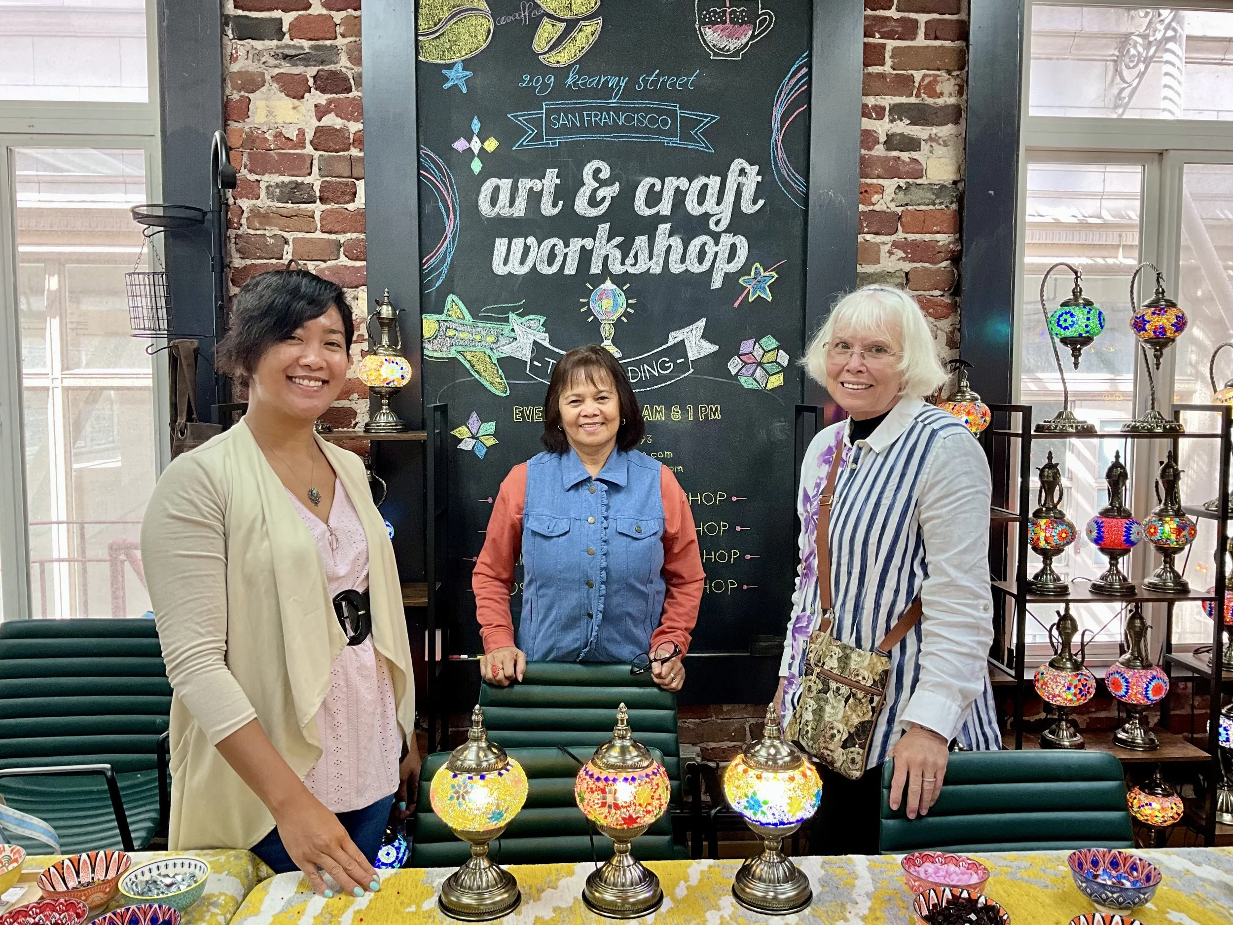 Three women standing behind a table with colorful mosaic lamps at an art and craft workshop in a room with brick walls and large windows.