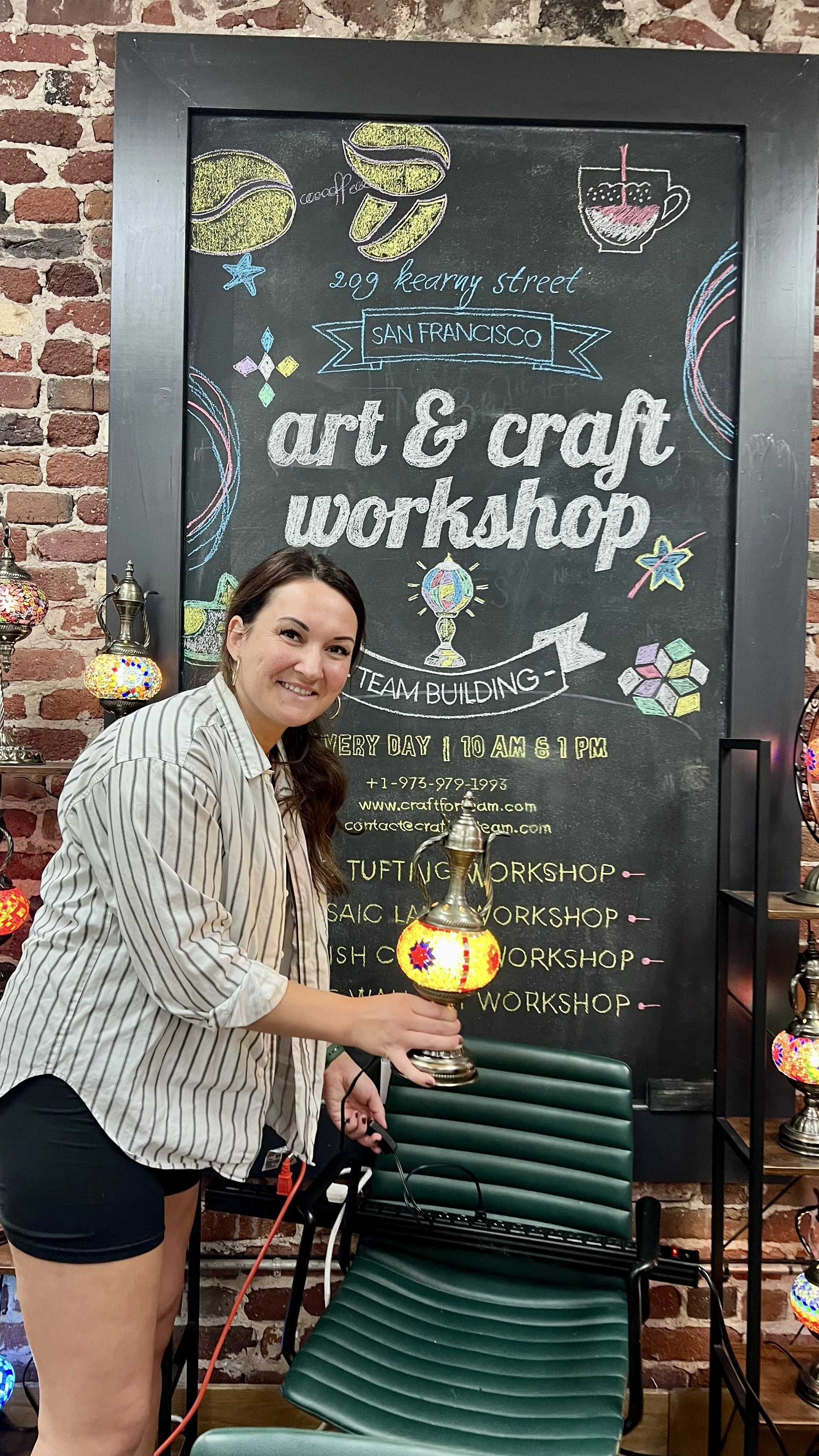 Woman smiling and holding a decorative lamp in front of a chalkboard sign advertising a chalk art and craft workshop in San Francisco.