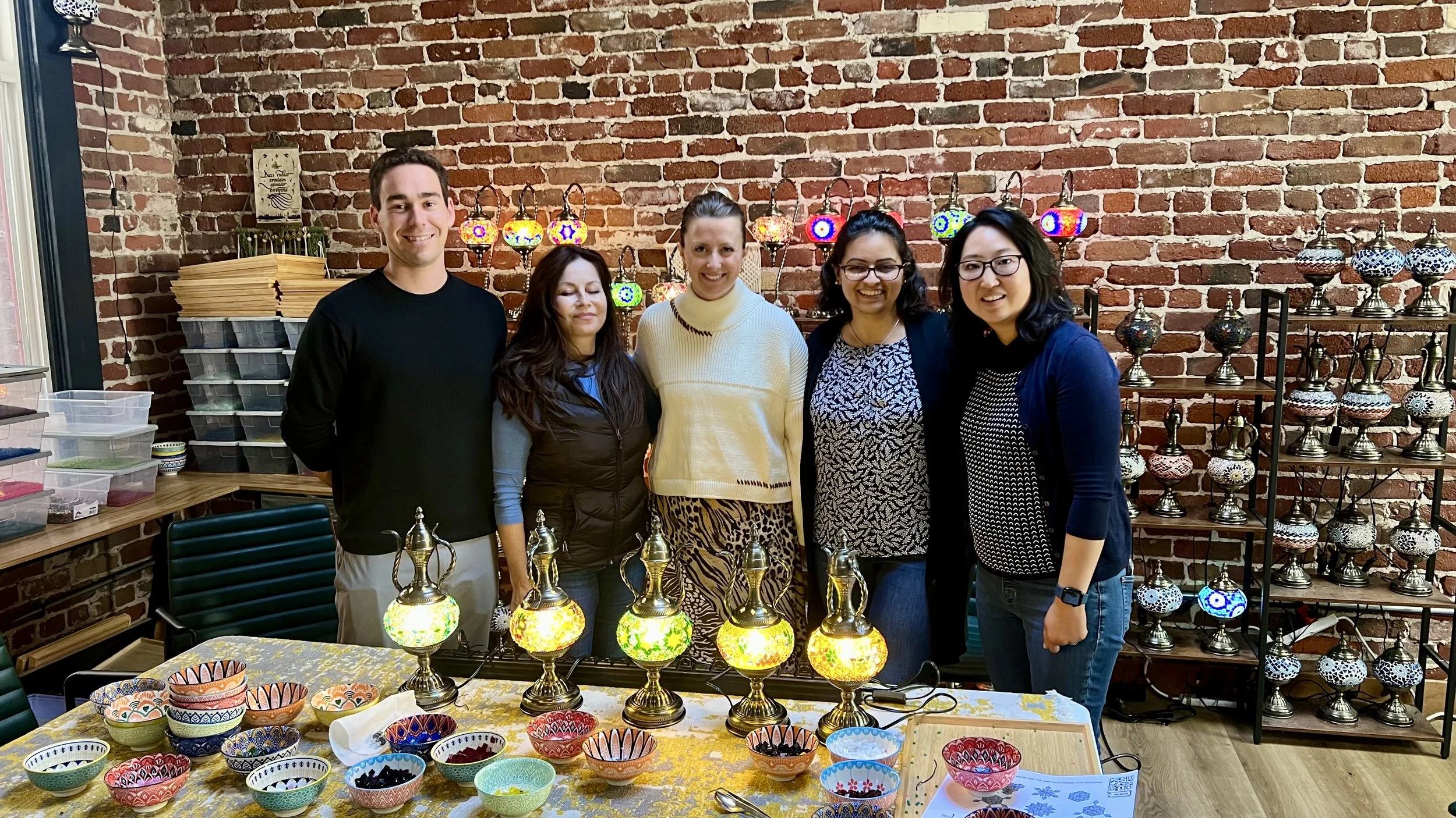 Five people standing behind a table with colorful lamps and bowls, in front of a brick wall and display shelves with decorated lamps.