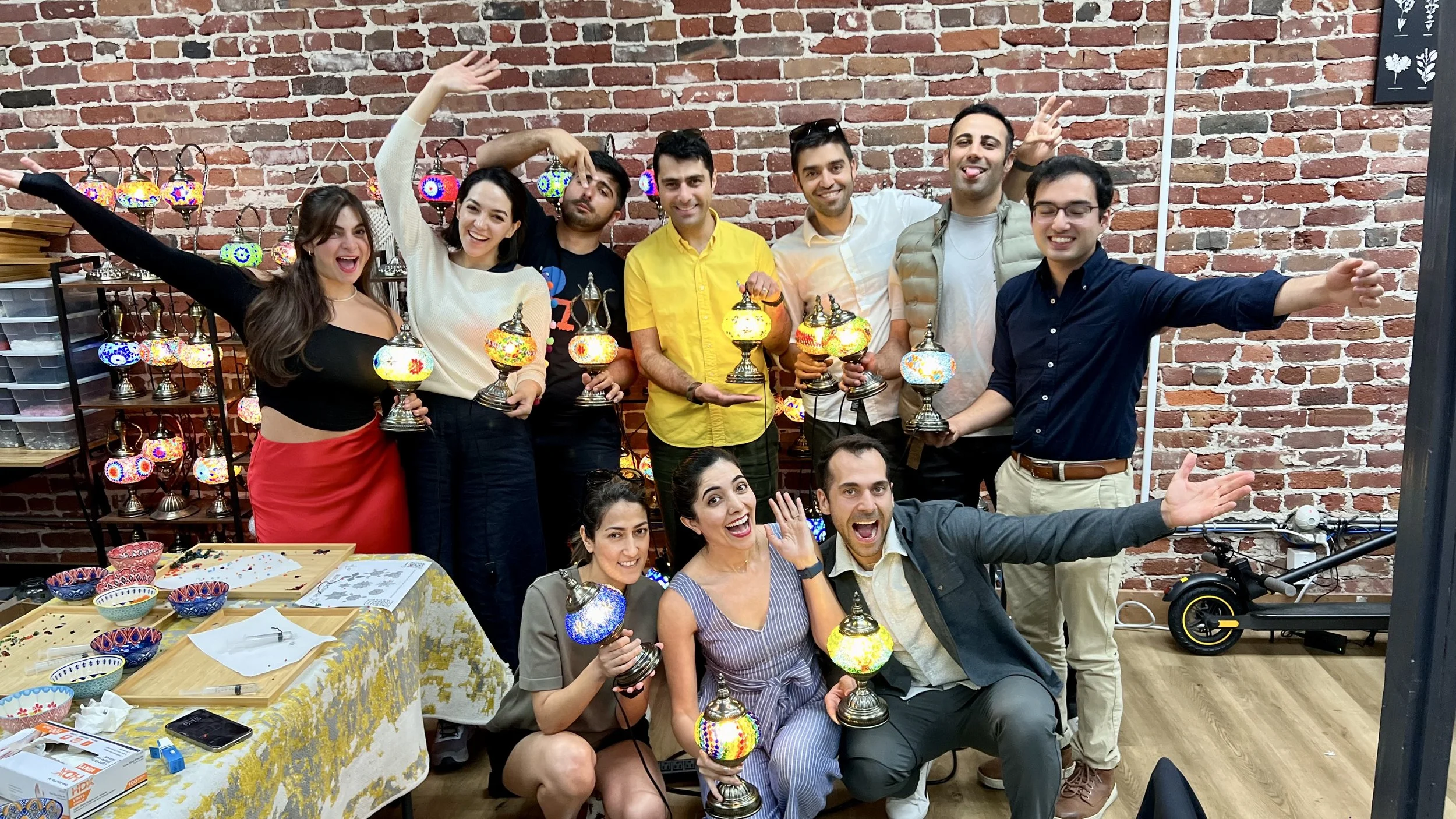 Group of nine people celebrating with colorful mosaic table lamps in a craft workshop, some holding lamps, others posing joyfully, in front of a brick wall decorated with more lamps.