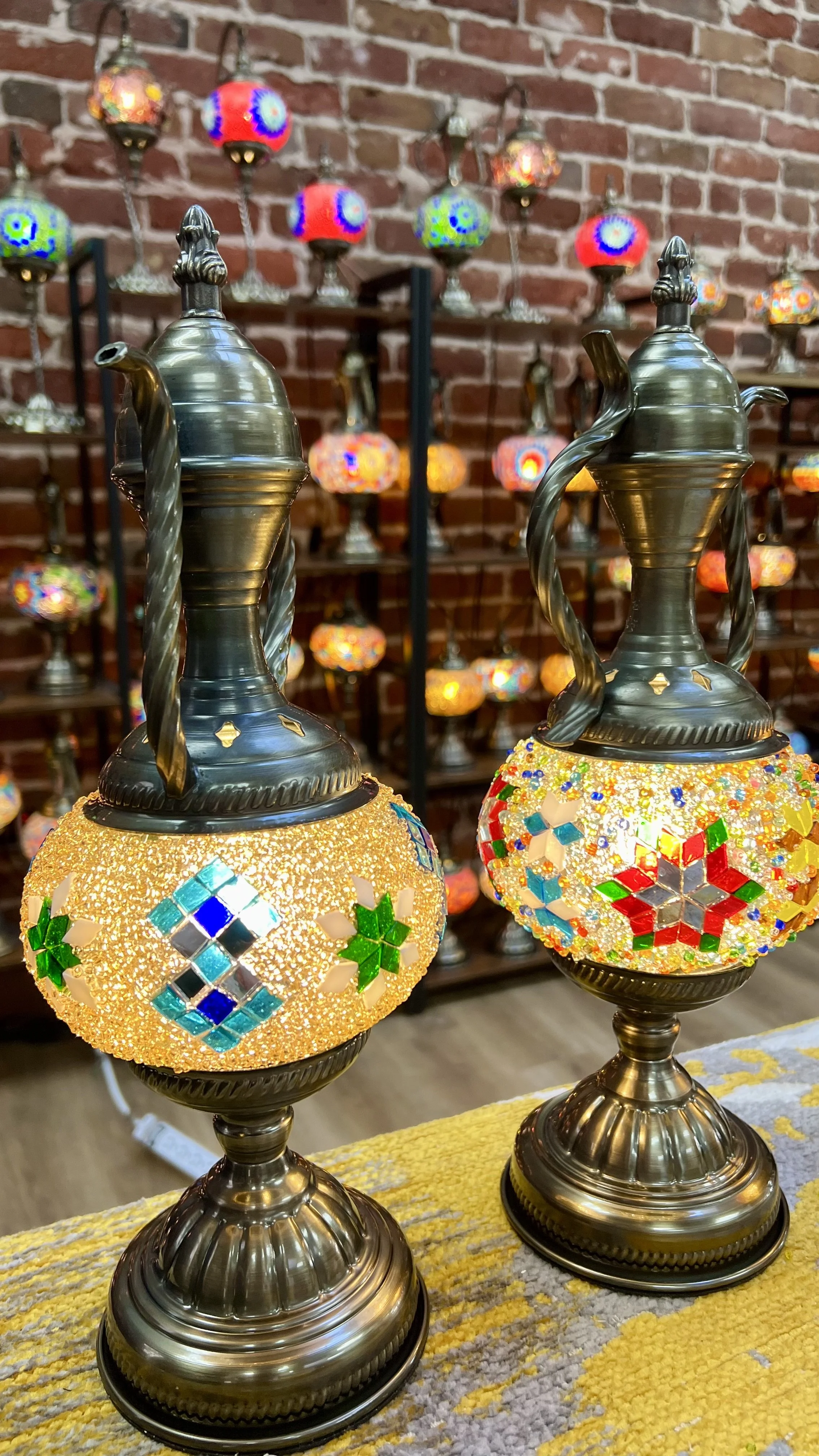 Two decorative mosaic lamps with metal bases and ornate metal tops, displayed on a table, with a background of a brick wall and a shelf filled with more colorful mosaic lamps.