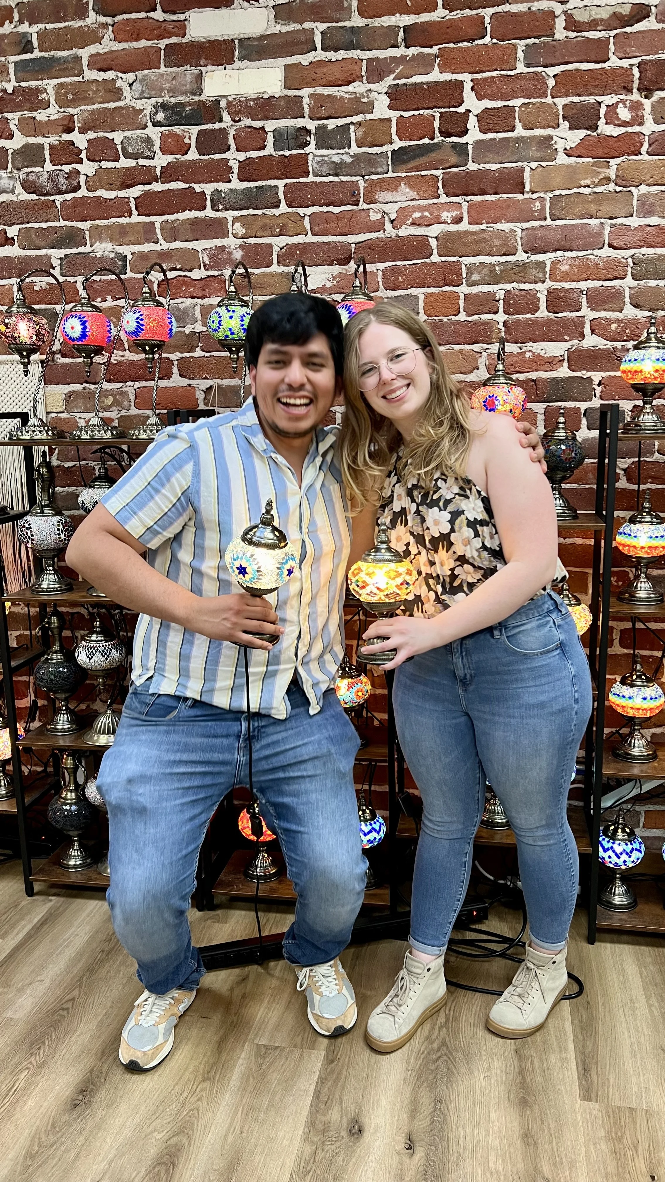 A young man and woman smiling and holding colorful mosaic lamps in front of a display of similar lamps against an exposed brick wall.