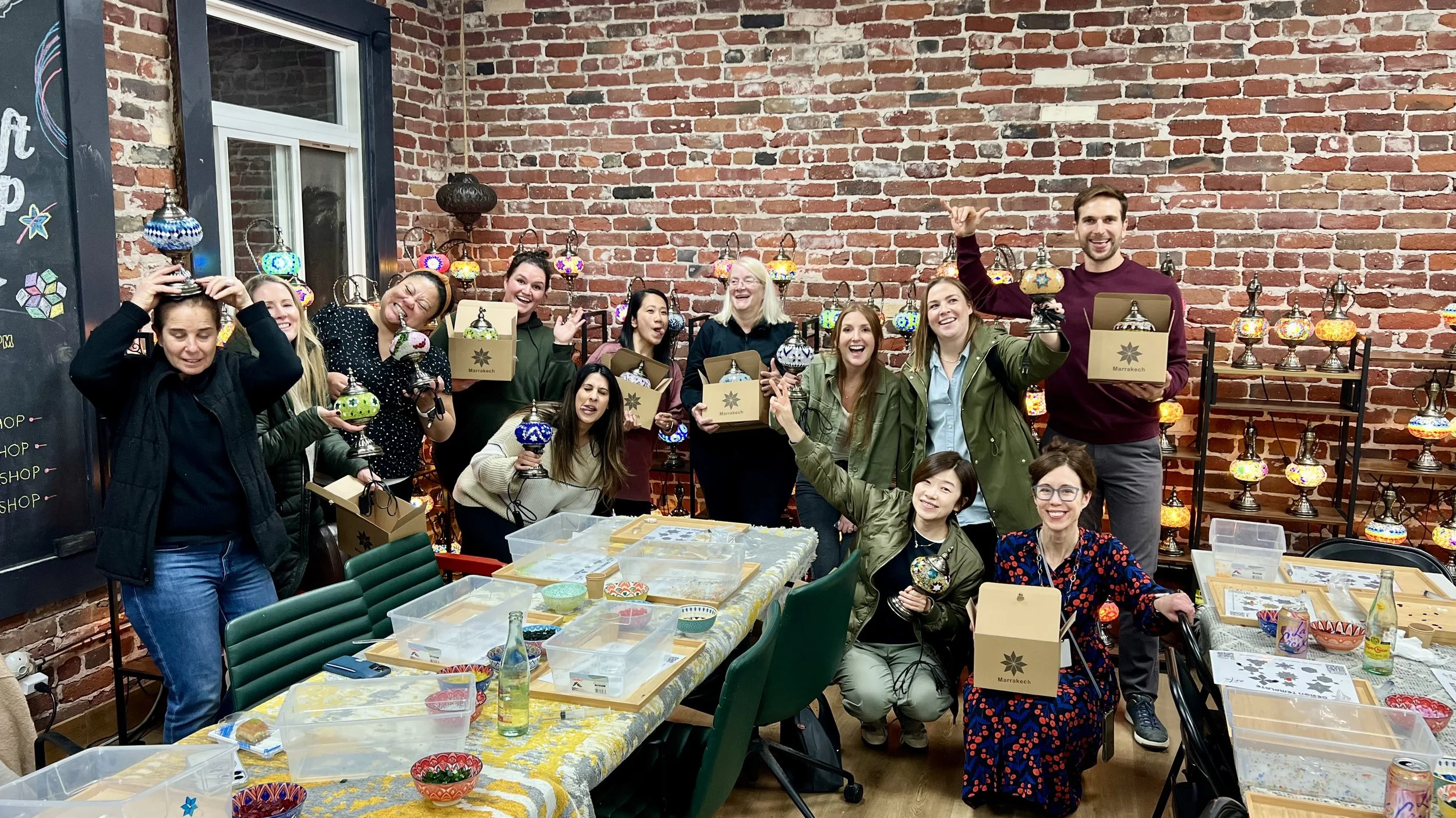 Group of people at a craft workshop holding up colorful decorative lanterns, with tables filled with craft supplies and a brick wall with hanging lanterns in the background.