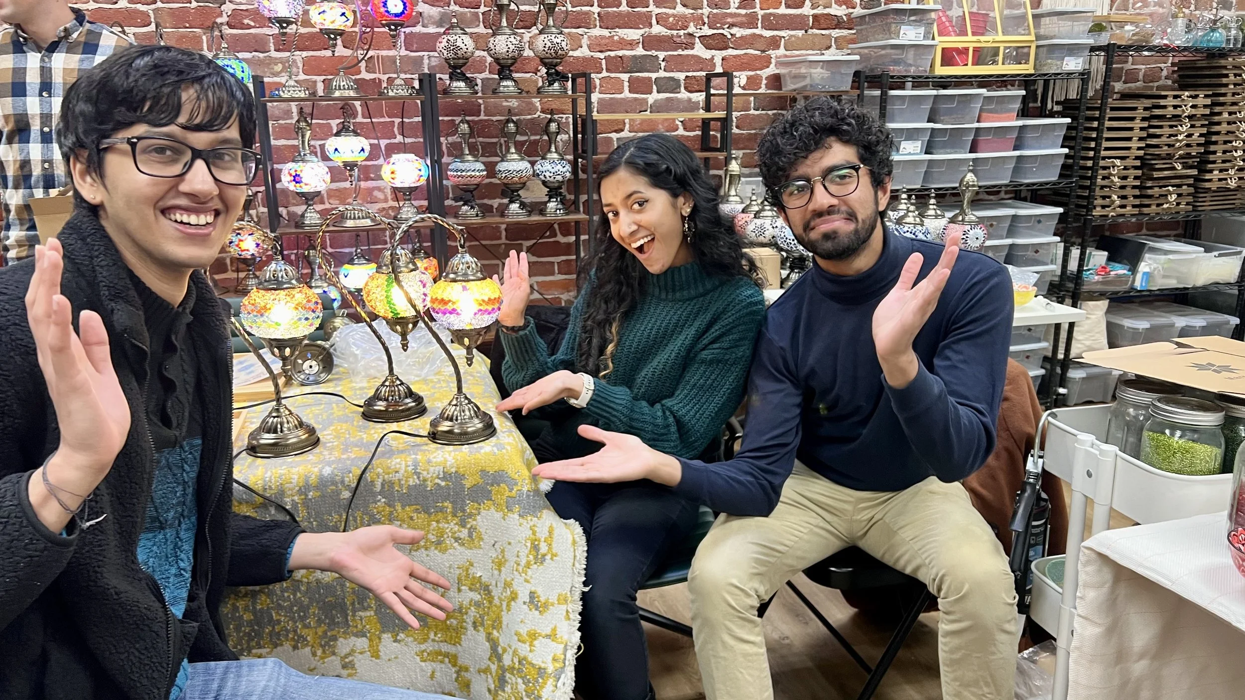 Three people sitting at a table displaying colorful decorative lamps, smiling and waving at the camera, with shelves of storage boxes and decorative items in the background.