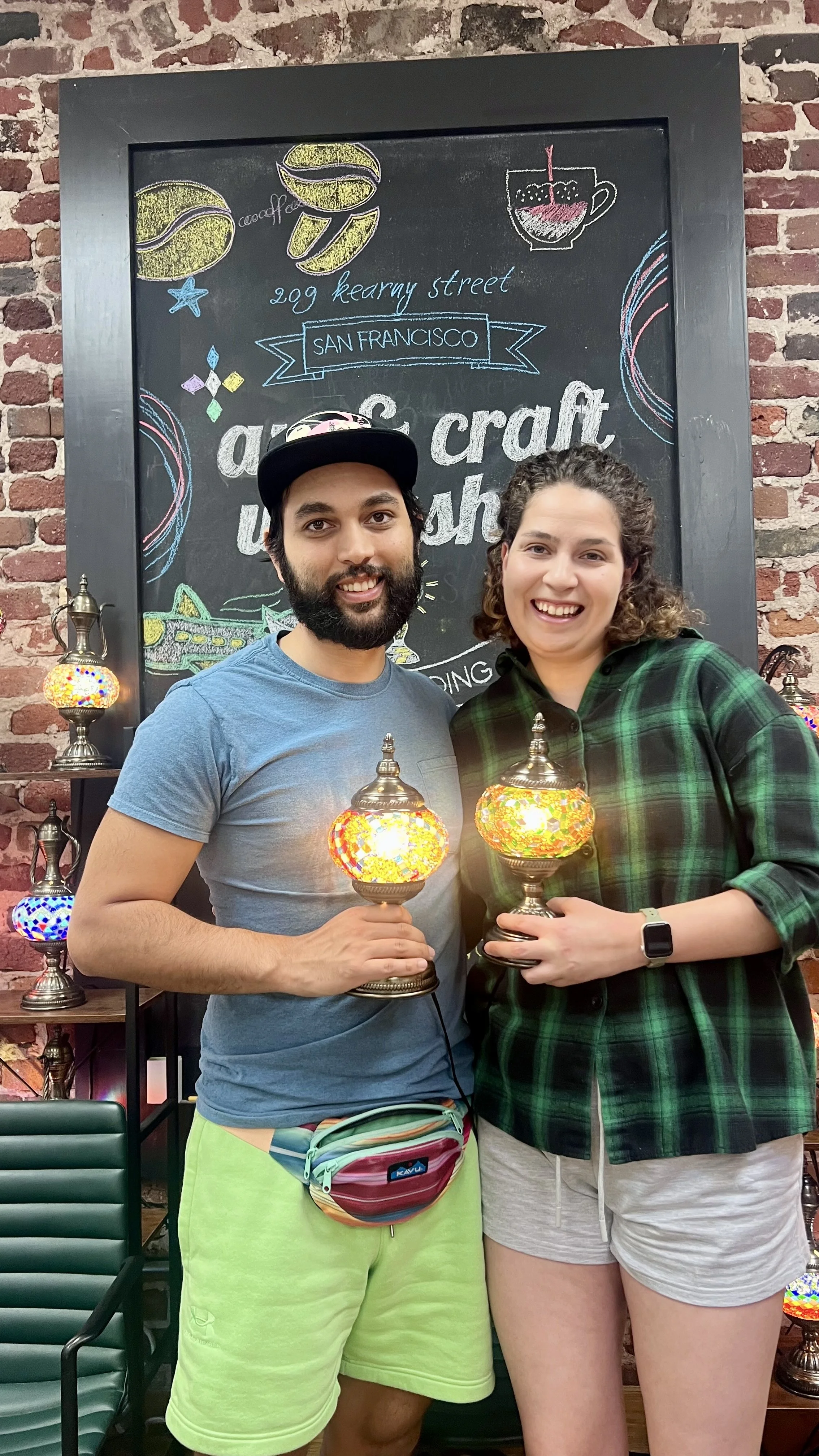 A man and woman smiling and holding colorful mosaic lamps inside a craft shop. The man is wearing a blue t-shirt, lime green shorts, a black cap, and a colorful fanny pack. The woman is wearing a green and black plaid shirt and light-colored shorts. 