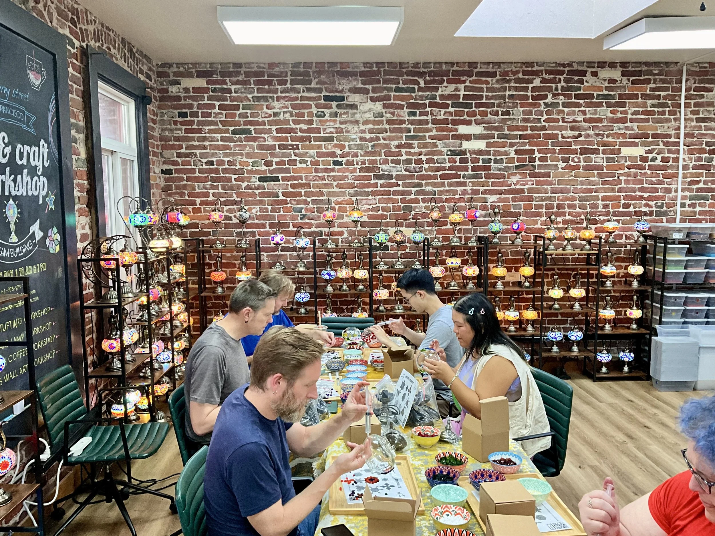 People participating in a mosaic craft workshop at a table filled with small bowls of colored glass pieces and mosaic patterns, with shelves of colorful mosaic lamps against a brick wall in the background.